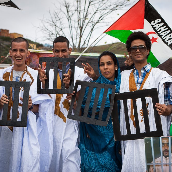 L'image montre un groupe de personnes posant ensemble lors d'une manifestation. Ils portent des vêtements traditionnels, notamment des robes blanches. Certains affichent des signes pacifiques avec leurs mains, tandis que d'autres tiennent des panneaux représentant des barreaux de prison. En arrière-plan, on peut voir un drapeau aux couleurs du Sahara. L'atmosphère semble engagée et solidaire.