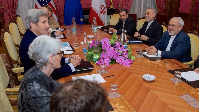 L'image montre une réunion diplomatique autour d'une grande table en bois. Des personnes sont assises de chaque côté, impliquées dans une discussion. On aperçoit des drapeaux d'États-Unis, de l'Union européenne et d'Iran sur la table. Au centre, il y a un bouquet de fleurs, probablement des orchidées. L'ambiance semble sérieuse mais productive, avec des participants au visage souriant, indiquant un échange positif.