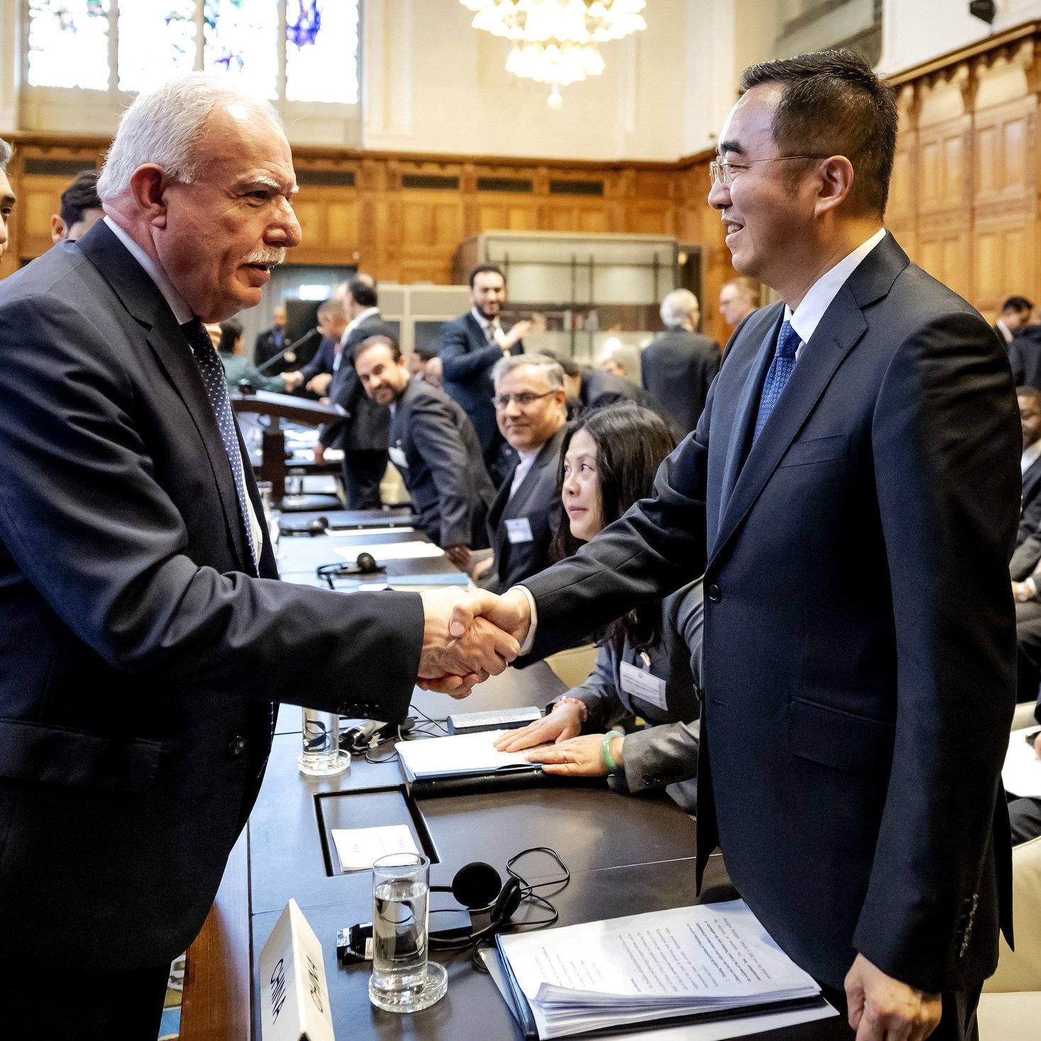 L'image montre une scène de réunion ou de conférence, probablement dans un cadre formel. Deux hommes se serrent la main, souriant, ce qui évoque une interaction chaleureuse ou un accord. En arrière-plan, on aperçoit d'autres participants qui semblent également engagés dans des discussions. La salle est spacieuse, avec une décoration élégante, des fenêtres laissant passer une lumière naturelle, et des sièges disposés autour d'une grande table. L'atmosphère semble professionnelle et collaborative.