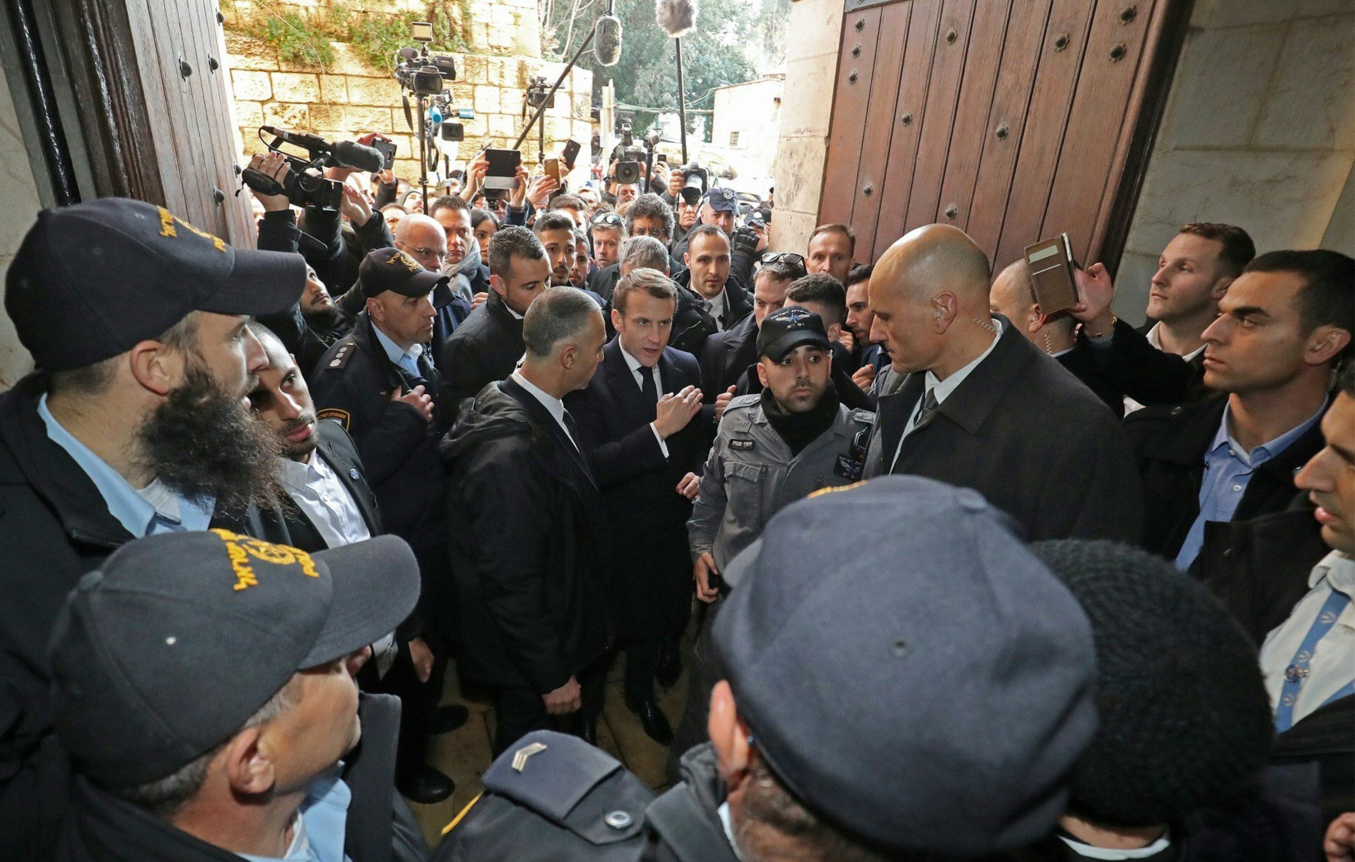 تصویر گروهی از افراد را نشان می‌دهد که در یک محیط شلوغ با امنیت زیاد در حال گفتگو هستند. تعدادی از افراد یونیفورم‌پوش به نظر می‌رسد که نگهبان هستند و در حال محافظت از یک فرد دیگر هستند. افراد دیگر در اطراف آن‌ها ایستاده‌اند و به نظر می‌رسد که برخی از آن‌ها مشغول ضبط یا عکاسی از این صحنه هستند. فضا به شدت پر ازدحام و حاکی از توجه رسانه‌ای است.