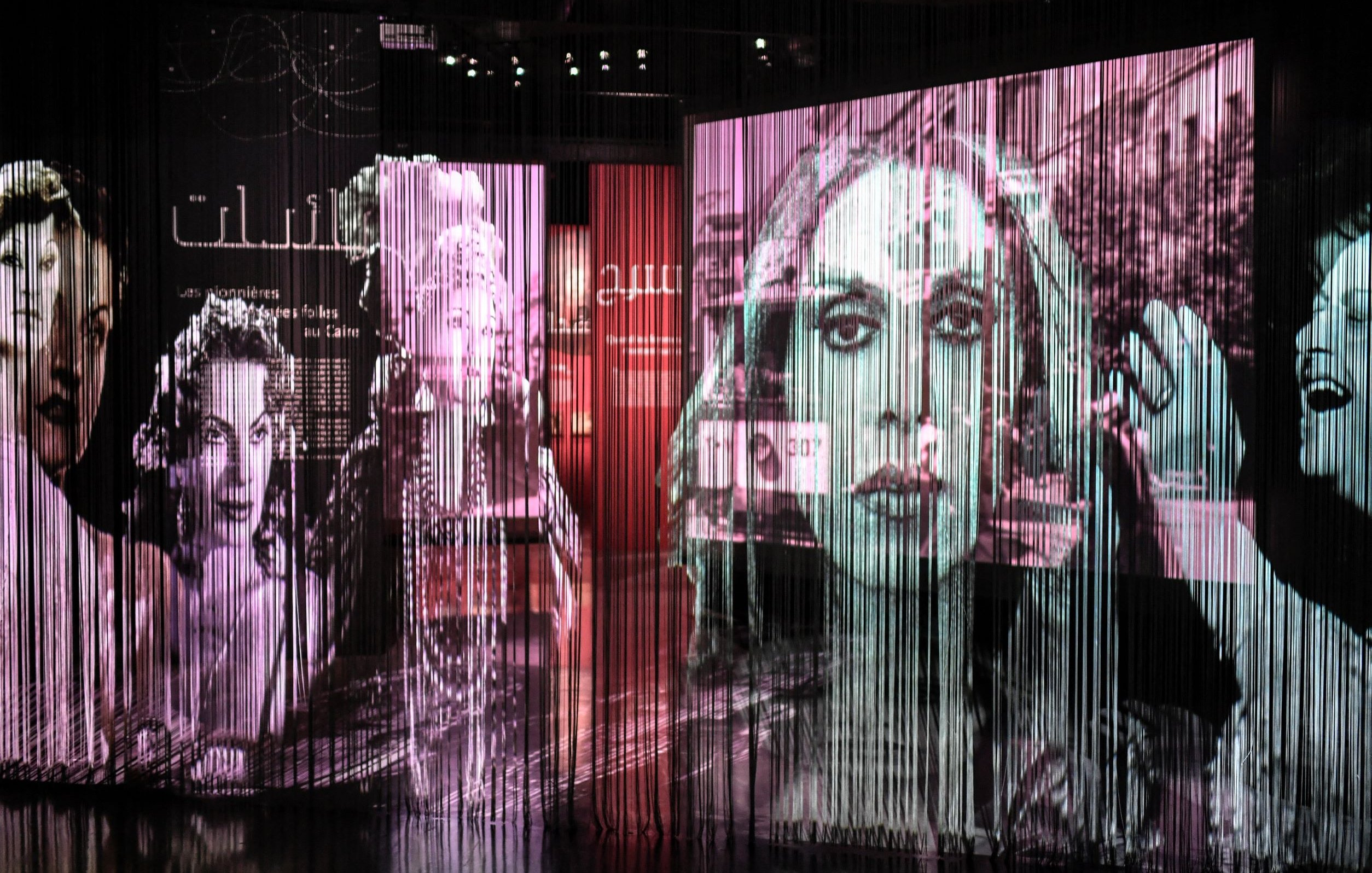 L'image présente une installation artistique composée de plusieurs grands panneaux transparents sur lesquels sont projetées des images en noir et blanc de visages féminins. Les projections créent un effet de profondeur et de mouvement, avec des formes et des silhouettes qui se superposent. Le fond est teinté de couleurs douces, suggérant une ambiance à la fois artistique et contemplative. Ce type d'installation évoque une réflexion sur la représentation et l'identité, tout en intégrant des éléments visuels captivants.