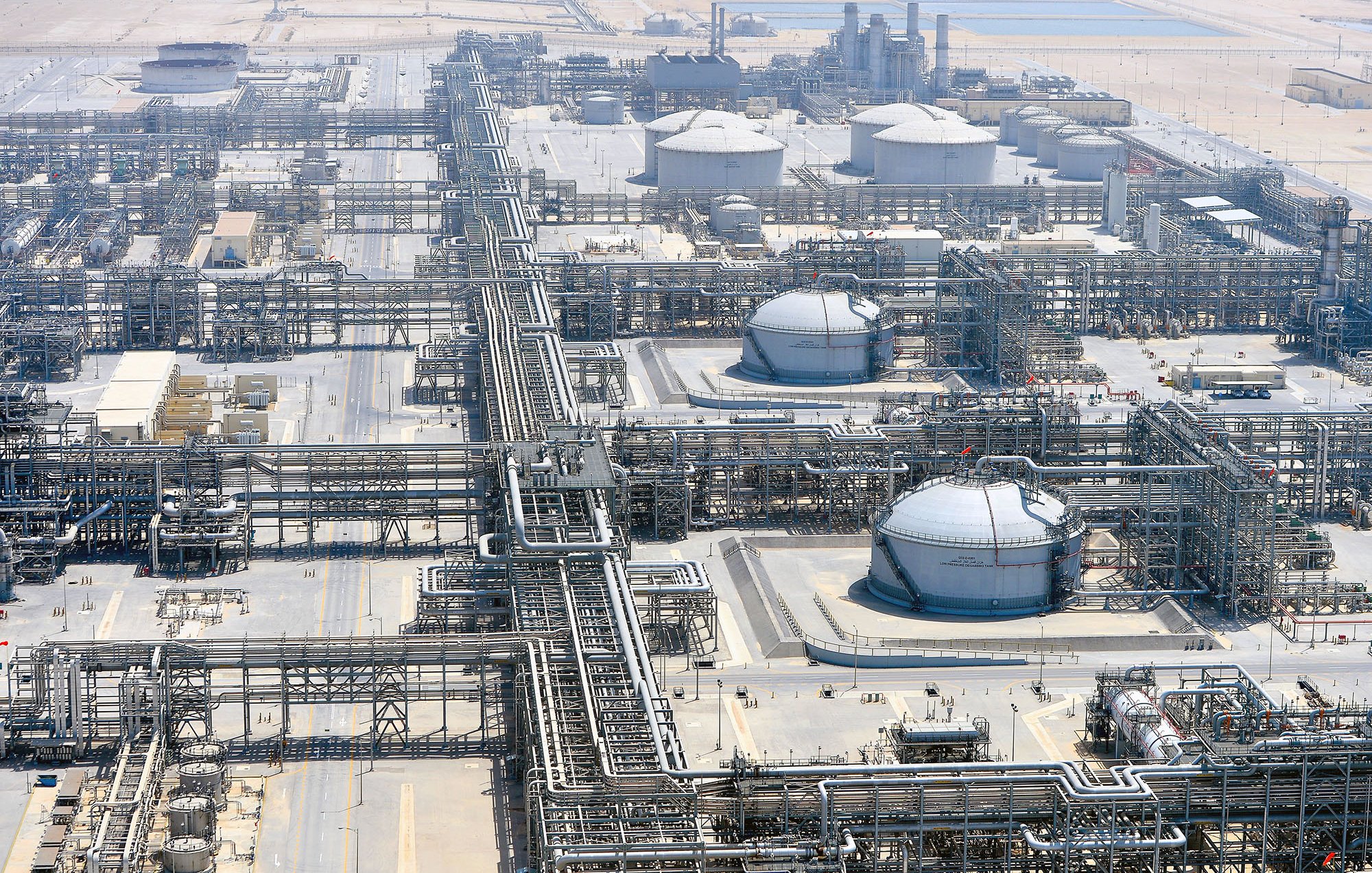 L'immagine mostra un impianto industriale, probabilmente una raffineria o un complesso di estrazione di prodotti petroliferi. Si possono vedere grandi serbatoi rotondi e una vasta rete di tubazioni che si snodano attraverso il sito. L'area circostante appare deserta, con una superficie di sabbia o terra arida. Sullo sfondo si intravedono strutture alte, che potrebbero essere torri di raffineria o impianti di trattamento. L'intero complesso è ben organizzato, con una disposizione sistematica delle tubazioni e dei serbatoi.