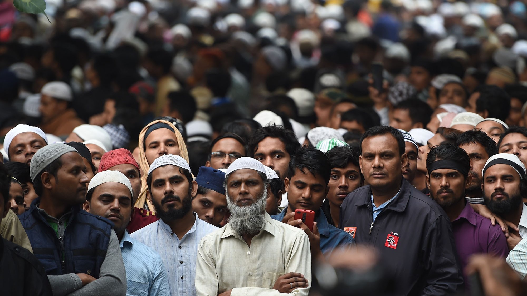 تظهر الصورة تجمعًا كبيرًا من الأشخاص، حيث يرتدي معظمهم القبعات البيضاء التقليدية، مما يدل على أنهم قد يكونون من أحد الثقافات الإسلامية. يظهر في مقدمة الصورة رجل ذو لحية رمادية، بينما تبدو وجوه الآخرين مملوءة بالتعبير عن الاهتمام والتركيز. الخلفية تشير إلى حشد كبير، مع تفاصيل غير واضحة بسبب كثافة الحضور. يبدو أن الجميع مجتمعون لمناسبة معينة أو حدث مهم.