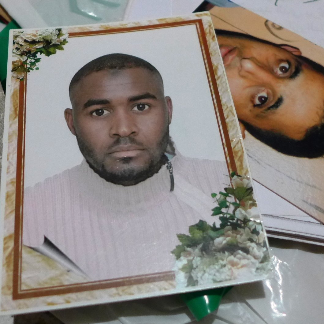 The image shows two photographs. One is a formal portrait of a man with short hair, wearing a light-colored sweater. The background contains decorative elements like flowers. The second photo appears to be a more candid or casual shot of another man, who has dark, curly hair and a neutral expression. Both photos are placed together on a surface that includes some papers and miscellaneous items.