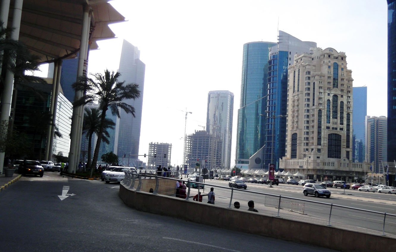 تظهر الصورة مشهداً حضرياً لمدينة حديثة، حيث تتداخل المباني الشاهقة مع تشكيلة متنوعة من التصاميم المعمارية. في الجهة اليسرى، توجد أشجار النخيل التي تضيف لمسة طبيعية على المشهد. يتواجد على الطريق مجموعة من السيارات، مما يشير إلى حركة المرور في المنطقة. السماء تظهر بلون فاتح، مما يوحي بأنها يوم مشمس. العمارات تتنوع في ارتفاعاتها وأشكالها، مما يعكس الطابع العصري للمدينة.