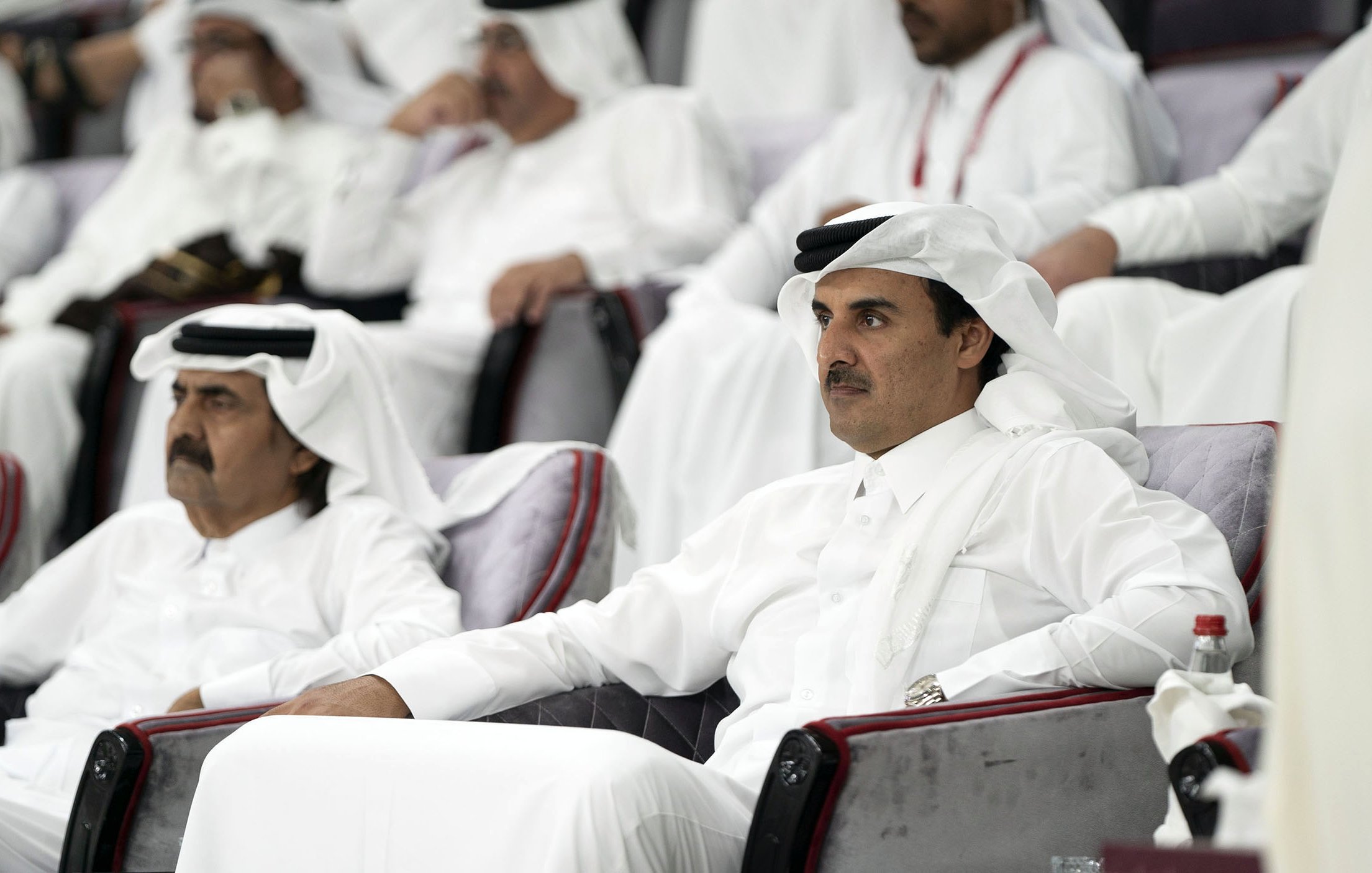 L'image montre un groupe de personnes assises dans des sièges, probablement dans un stade ou un lieu événementiel. Deux hommes sont au premier plan, vêtus de vêtements traditionnels qataris, avec des turbans blancs et des robes longues. Ils semblent attentifs et engagés dans l'événement se déroulant devant eux. L'atmosphère est celle d'un rassemblement formel, avec d'autres personnes en arrière-plan également habillées de manière traditionnelle.