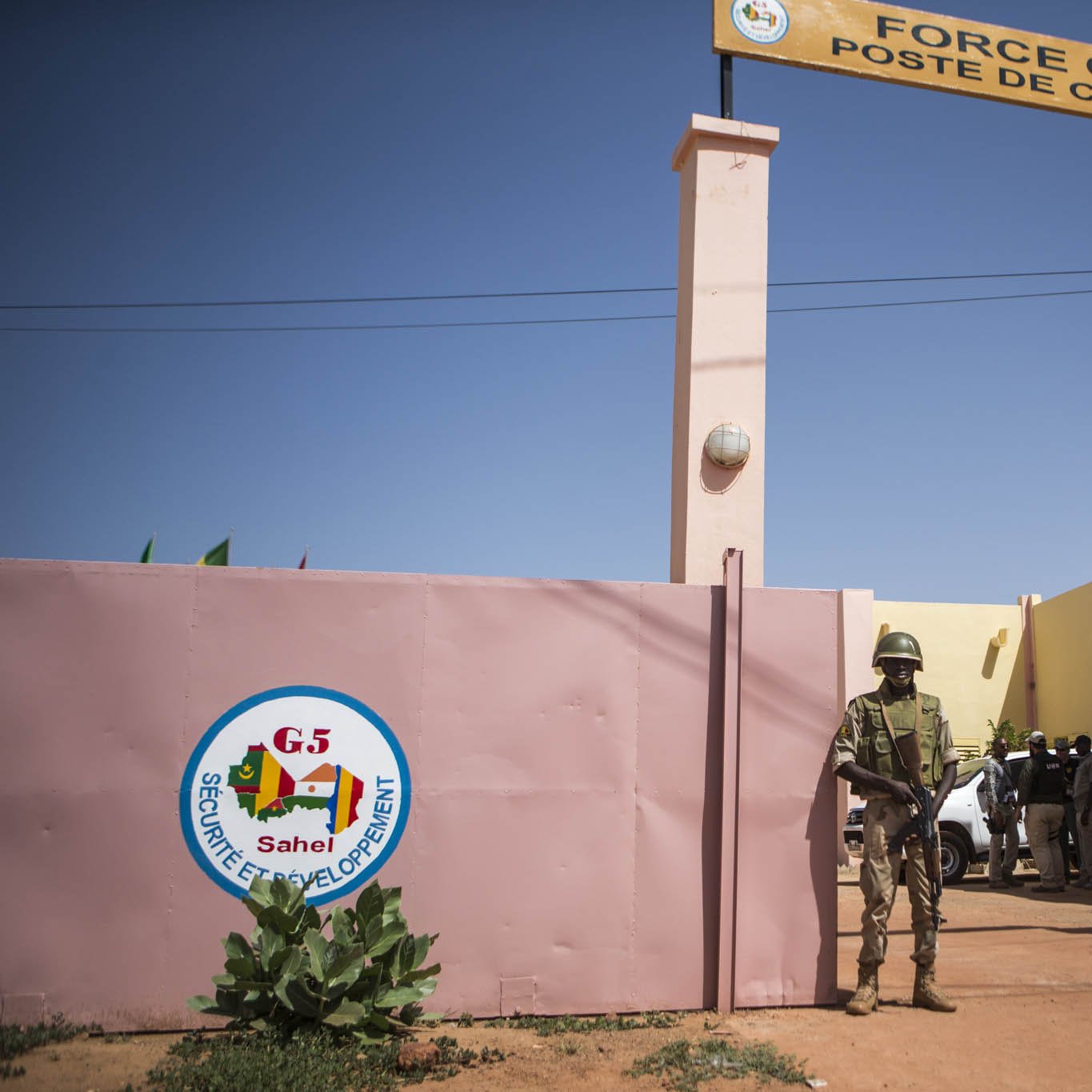 L'image montre un bâtiment qui semble être un poste de commandement de la "Force Conjointe" au sein du G5 Sahel. À l'entrée, on voit un mur avec le logo du G5, ainsi qu'un panneau indiquant "FORCE CONJOINTE POSTE DE COMMANDEMENT". Un soldat en uniforme est en position de garde près de l'entrée, tandis que d'autres personnes sont visibles à l'intérieur ou autour du bâtiment. L'environnement est désertique, et le ciel est dégagé.