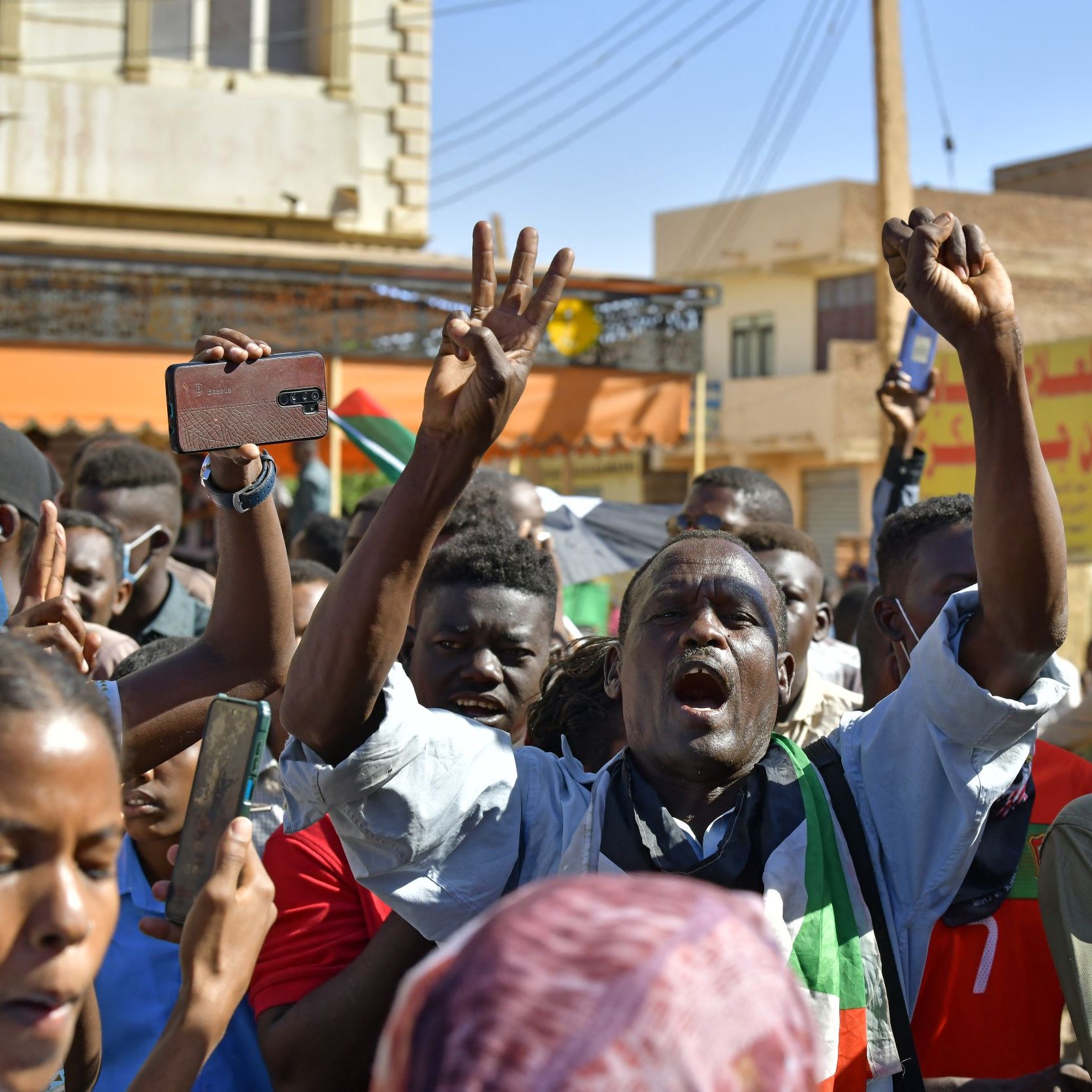 L'immagine mostra un gruppo di persone in una manifestazione vibrante. Gli individui sembrano esprimere passione e determinazione mentre sollevano le braccia e alcuni gridano slogan. Alcuni di loro indossano colori che richiamano una bandiera, suggerendo un forte attaccamento a una causa o a un movimento. È visibile anche un megafono, che indica che ci sono leader che cercano di comunicare con la folla. Lo sfondo presenta edifici e una scena urbana tipica di un contesto di protesta. L'atmosfera sembra intensa e carica di emozioni.