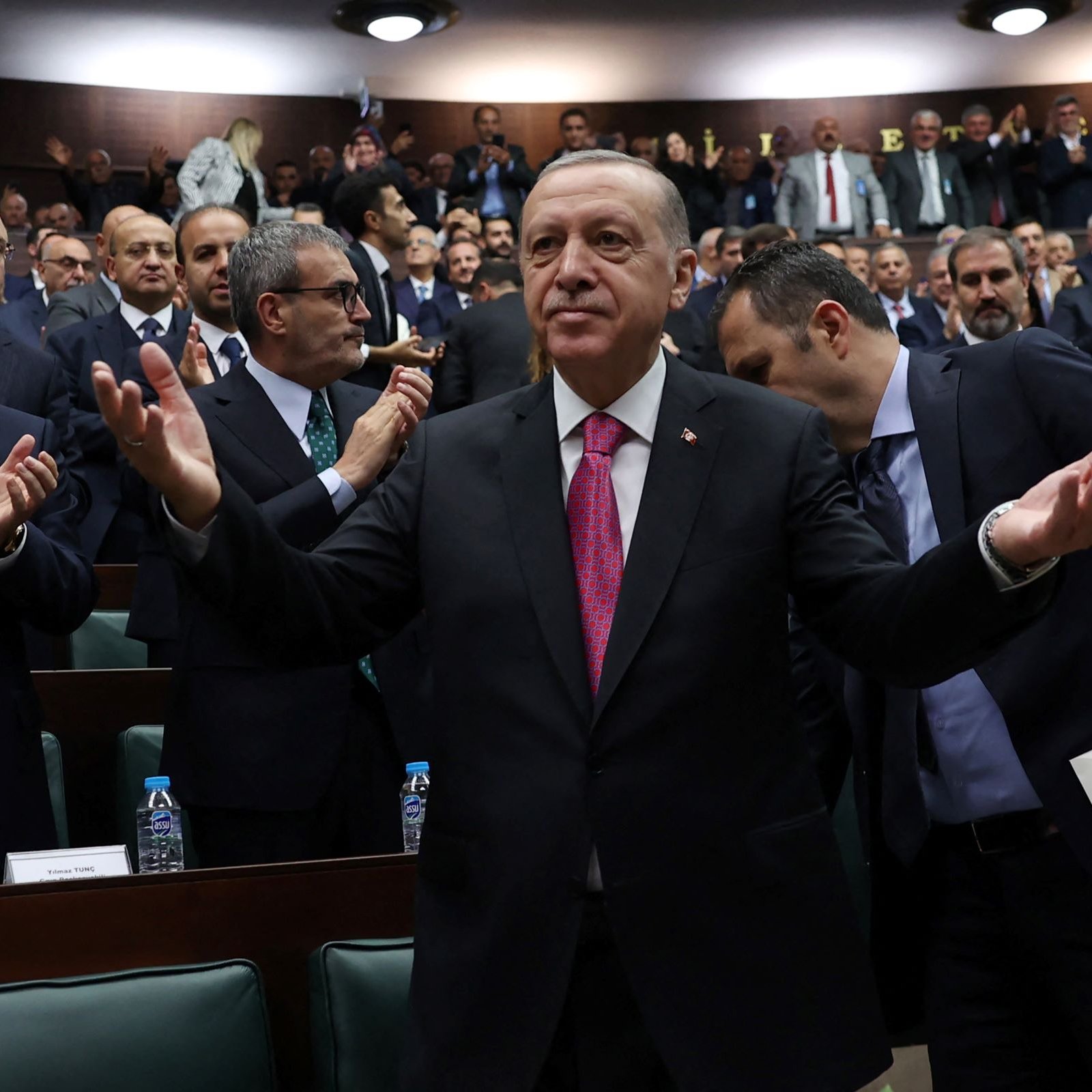 الصورة تظهر رئيس تركيا، رجب طيب أردوغان، standing في مجلس يتكون من عدد كبير من الشخصيات. يبدو أنه يتحدث أو يقدم خطابًا، حيث يرفع ذراعيه بشكل يعبر عن التواصل أو الاستجابة. خلفه، يوجد مجموعة من الحضور الذين يصفقون أو يستمعون له، مما يوحي بأجواء من التفاعل والحماس. ترتدي الشخصيات خلفه ملابس رسمية، ويبدو أنهم في جلسة برلمانية أو حدث سياسي.