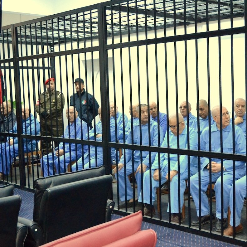 L'image montre un groupe d'individus assis derrière des barreaux. Ils portent tous des vêtements similaires, de couleur bleue. L'atmosphère semble sérieuse et formelle, peut-être en lien avec une audience ou un procès. Plusieurs personnes en uniforme sont présentes, suggérant un contexte de sécurité ou d'autorité. Les individus semblent concentrés ou préoccupés.