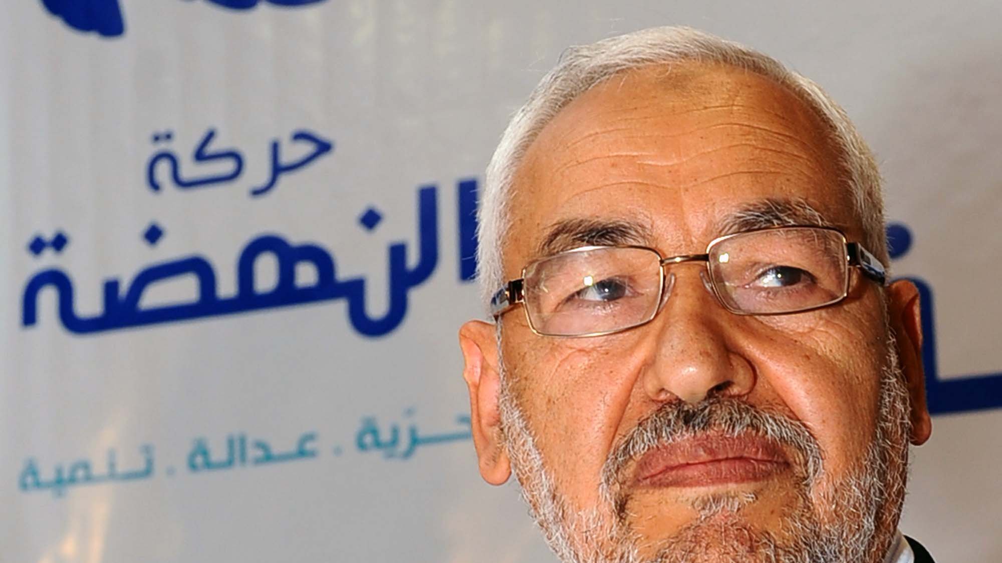 The image features a man with a gray beard and glasses, looking thoughtfully to the side. He is dressed in a light-colored shirt and blazer. In the background, there is a logo with Arabic text and a star, indicating it is related to a political movement or organization. The overall tone of the image suggests a serious or contemplative mood.