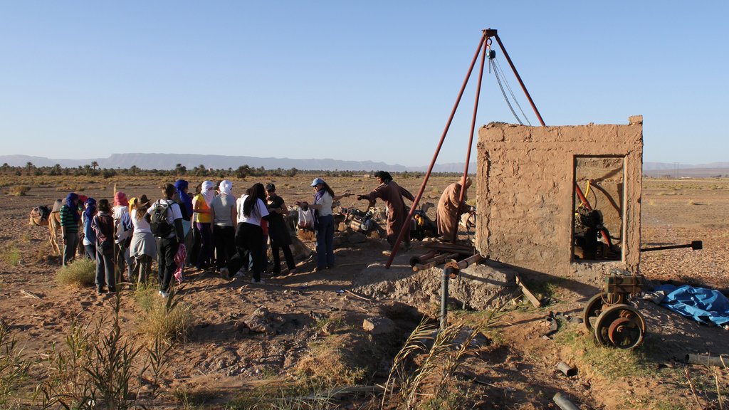 L'image montre un groupe de personnes rassemblées autour d'une structure en terre, possiblement une ancienne construction ou un puits. Il y a une machine ou un mécanisme visible à l'intérieur de la structure. L'environnement est désertique, avec un sol aride et des montagnes en arrière-plan. Le ciel est dégagé, reflétant une ambiance ensoleillée. Les participants semblent s'intéresser à l'activité en cours, observant attentivement.