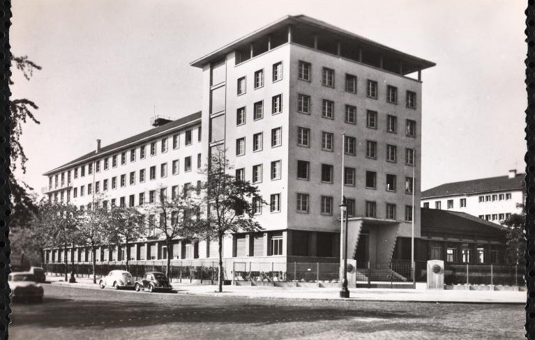 L'image représente un bâtiment architectural de plusieurs étages, probablement de style moderne. Il est photographié sous une lumière naturelle, mettant en avant sa façade aux lignes géométriques. Le bâtiment semble être entouré d'arbres et de quelques véhicules anciens, ce qui donne une ambiance d'époque. On peut également apercevoir des éléments de la rue, comme des lampadaires et éventuellement d'autres bâtiments en arrière-plan. L'ensemble de l'image présente une esthétique monochrome, accentuant le caractère historique de la scène.