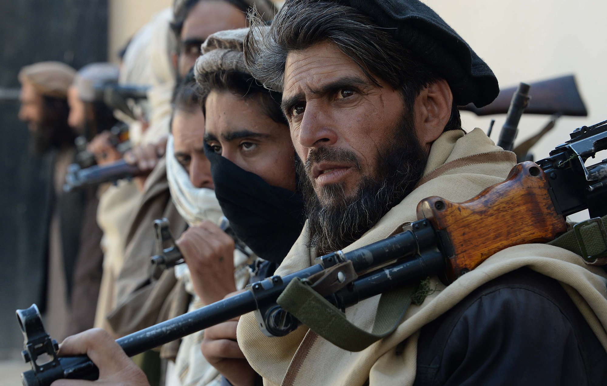 The image features a group of individuals standing closely together, each holding firearms. They appear to be dressed in traditional clothing, with some wearing headscarves and others with their faces partially obscured. The focus is on one man in the foreground, who has a serious expression and a well-groomed beard. The background is unfocused, but it suggests a rugged, possibly desert-like environment. The overall mood conveys tension or readiness.