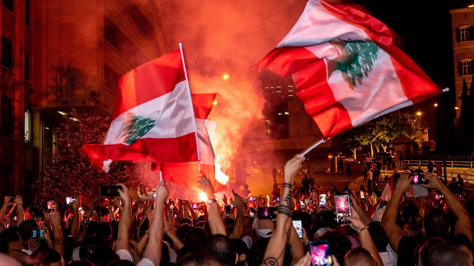تظهر الصورة تجمعًا حاشدًا في أحد الشوارع، حيث يحمل المتظاهرون أعلام لبنان. تضيء الأجواء بألوان النيران والدخان، مما يخلق جوًا احتفاليًا أو تعبيريًا. يُلاحظ أن الكثير من الأشخاص يستخدمون هواتفهم لتسجيل اللحظة أو التقاط الصور، بالإضافة إلى الأضواء الساطعة التي تضيف حيوية للمشهد.