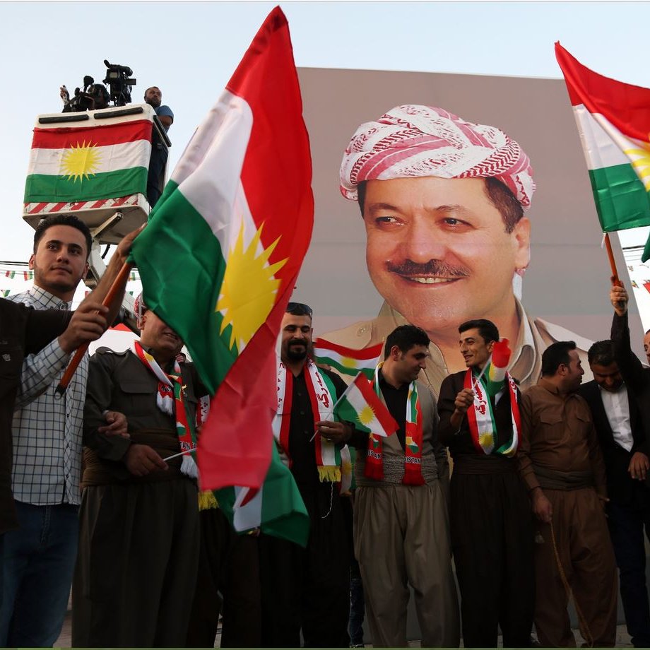 در تصویر گروهی از مردان لباس کردی به همراه پرچم‌های کردستان را مشاهده می‌کنیم. آن‌ها در حال ایستادن در مقابل یک بنر بزرگ هستند که تصویر فردی را نمایش می‌دهد. افراد با نشاط و خوشحالی پرچم‌ها را در دست دارند و به نظر می‌رسد که در یک مراسم یا جشن خاص شرکت کرده‌اند. فضای جمعی و پر انرژی به خوبی در تصویر حس می‌شود.