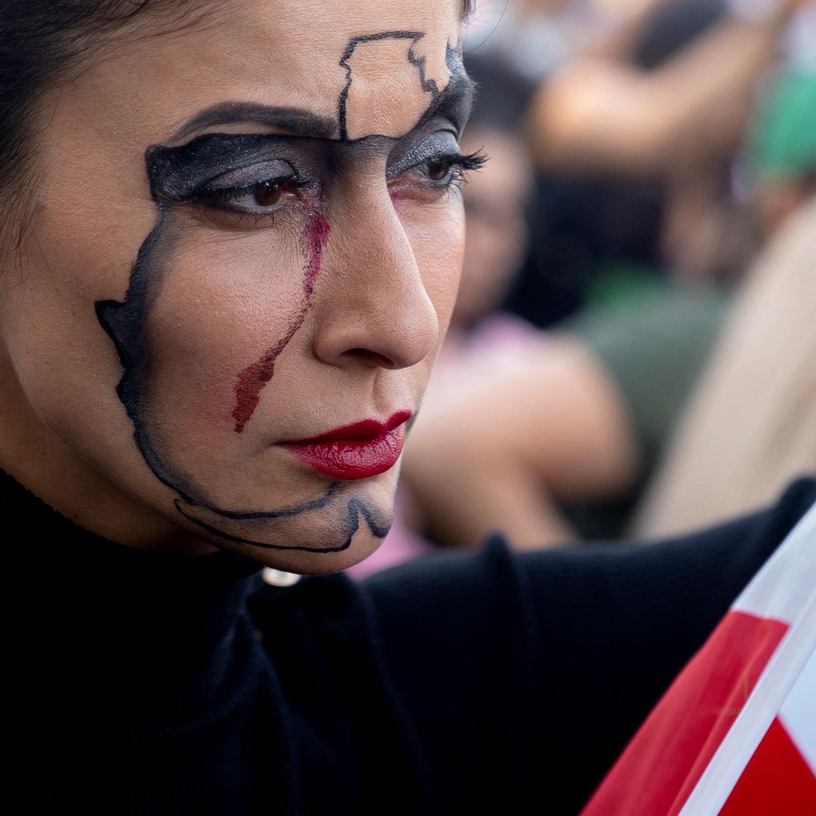 في هذه الصورة، تظهر امرأة تحمل علمًا بألوان الأخضر والأبيض والأحمر. تضع مكياجًا فنيًا على وجهها يتضمن خطوطًا سوداء ودموعًا باللون الأحمر. تعبير وجهها جاد، والمشهد يبدو مليئًا بالناس في الخلفية. الجو العام يشير إلى أن هناك فعاليات أو أحداث تجري في المكان.