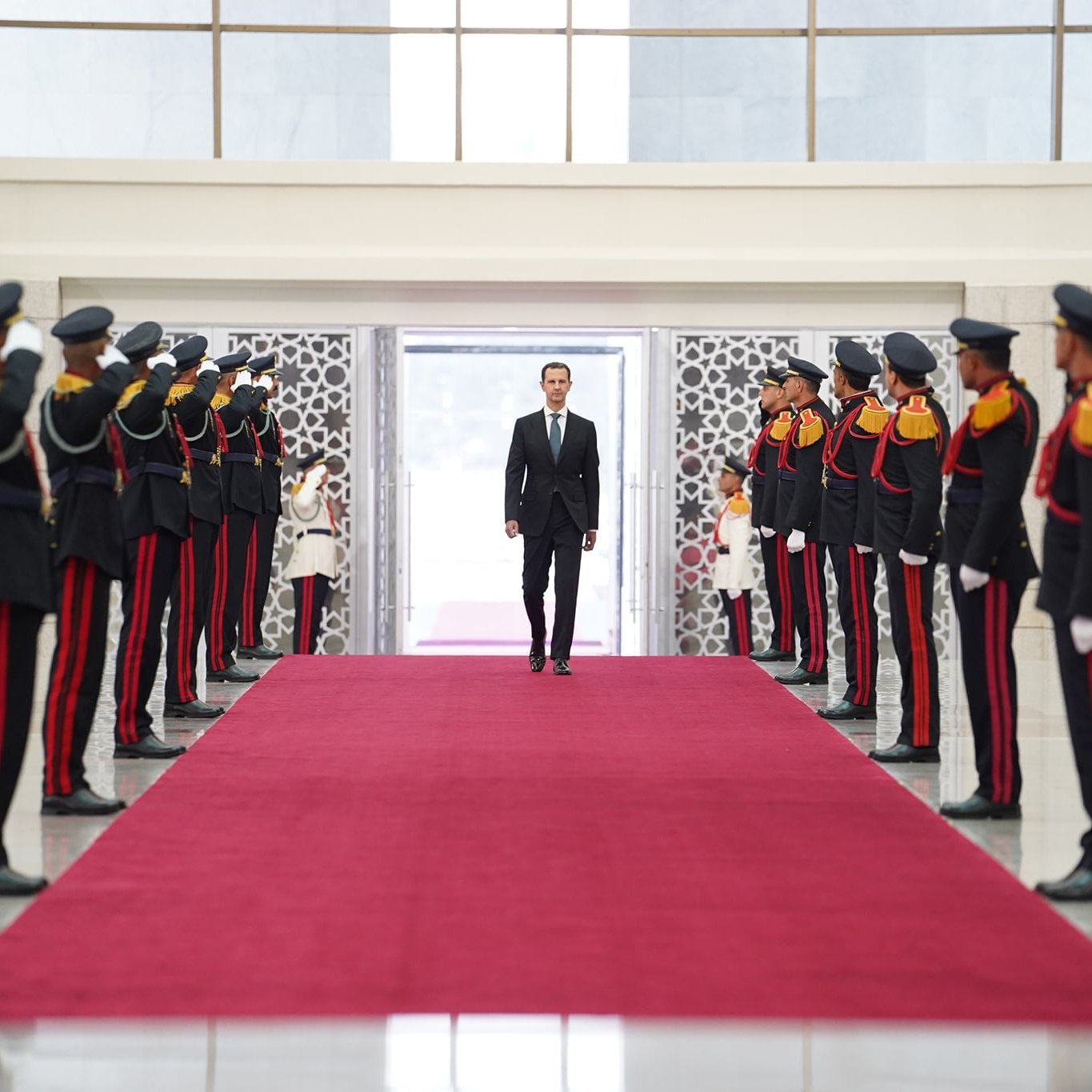 En la imagen se observa un pasillo alfombrado de rojo, flanqueado por un grupo de soldados en uniforme, que están en posición de saludo. Al fondo, se puede ver a un hombre vistiendo un traje negro que camina hacia adelante. El ambiente parece ser formal y está bien iluminado, con paredes de mármol y grandes ventanas que permiten la entrada de luz natural. El contexto sugiere un evento oficial o ceremonia.