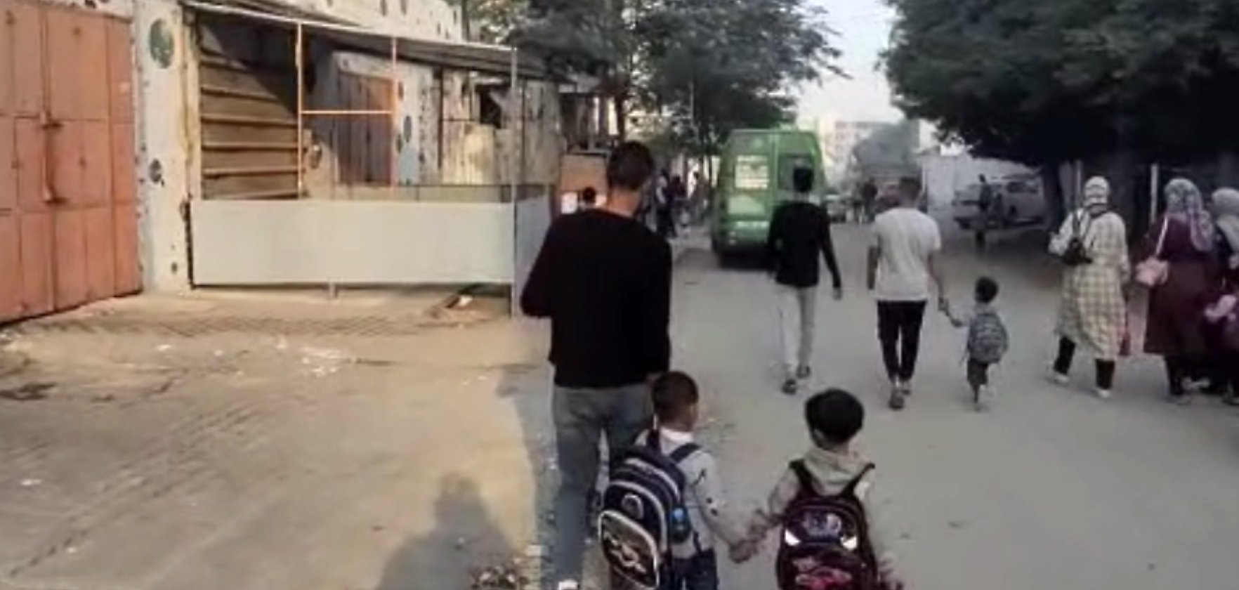 Un uomo cammina con due bambini, mentre altre persone si muovono in una strada cittadina.