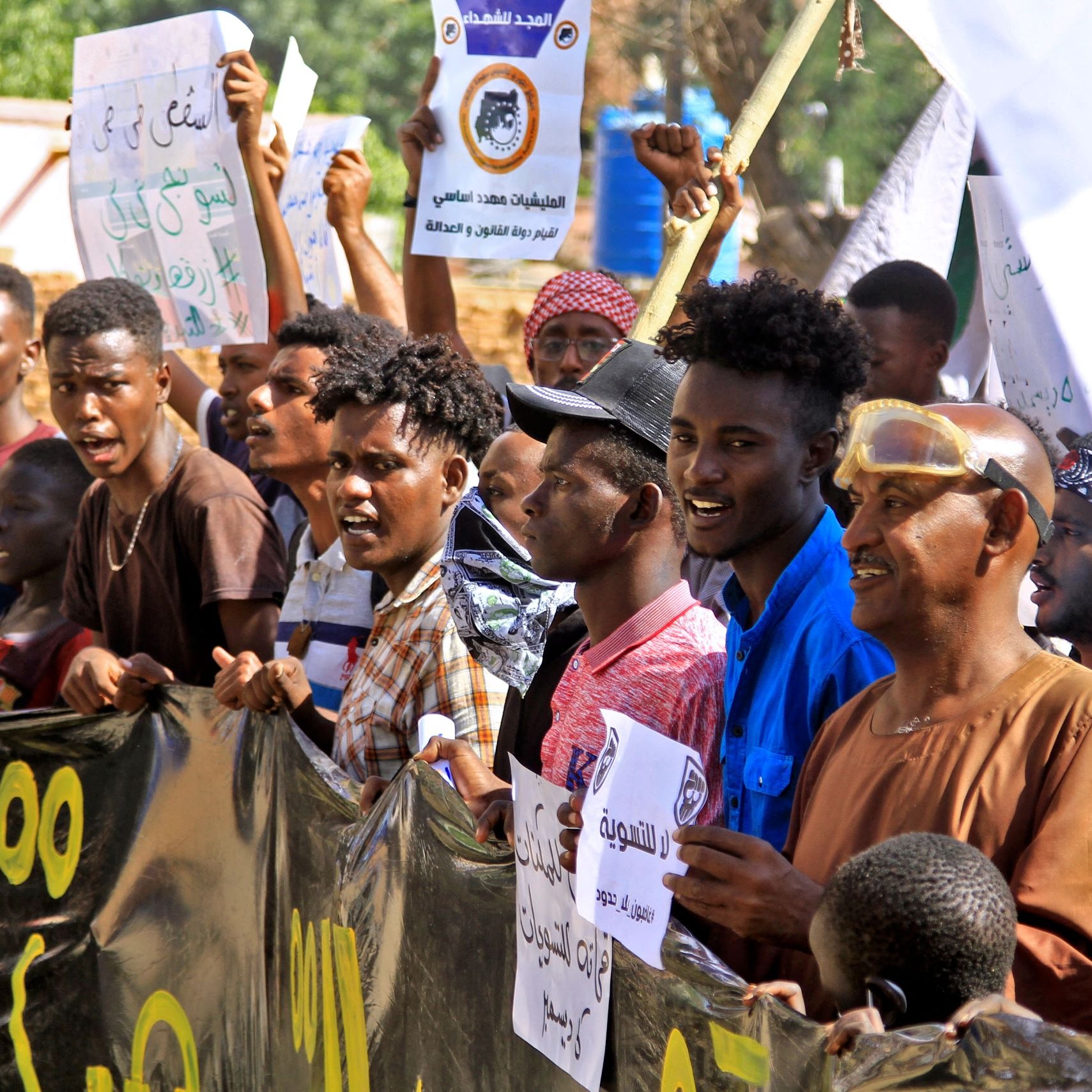 الصورة تظهر مجموعة من الناس في تظاهرة، يحملون لافتات و شعارات. يبدو أنهم متحمسون ويعبرون عن آرائهم. يظهر في الخلفية بعض المباني والأشجار، مما يدل على أن التجمع يحدث في منطقة مفتوحة. الوجوه تعكس مشاعر مختلفة، مثل الحماس والعزيمة.