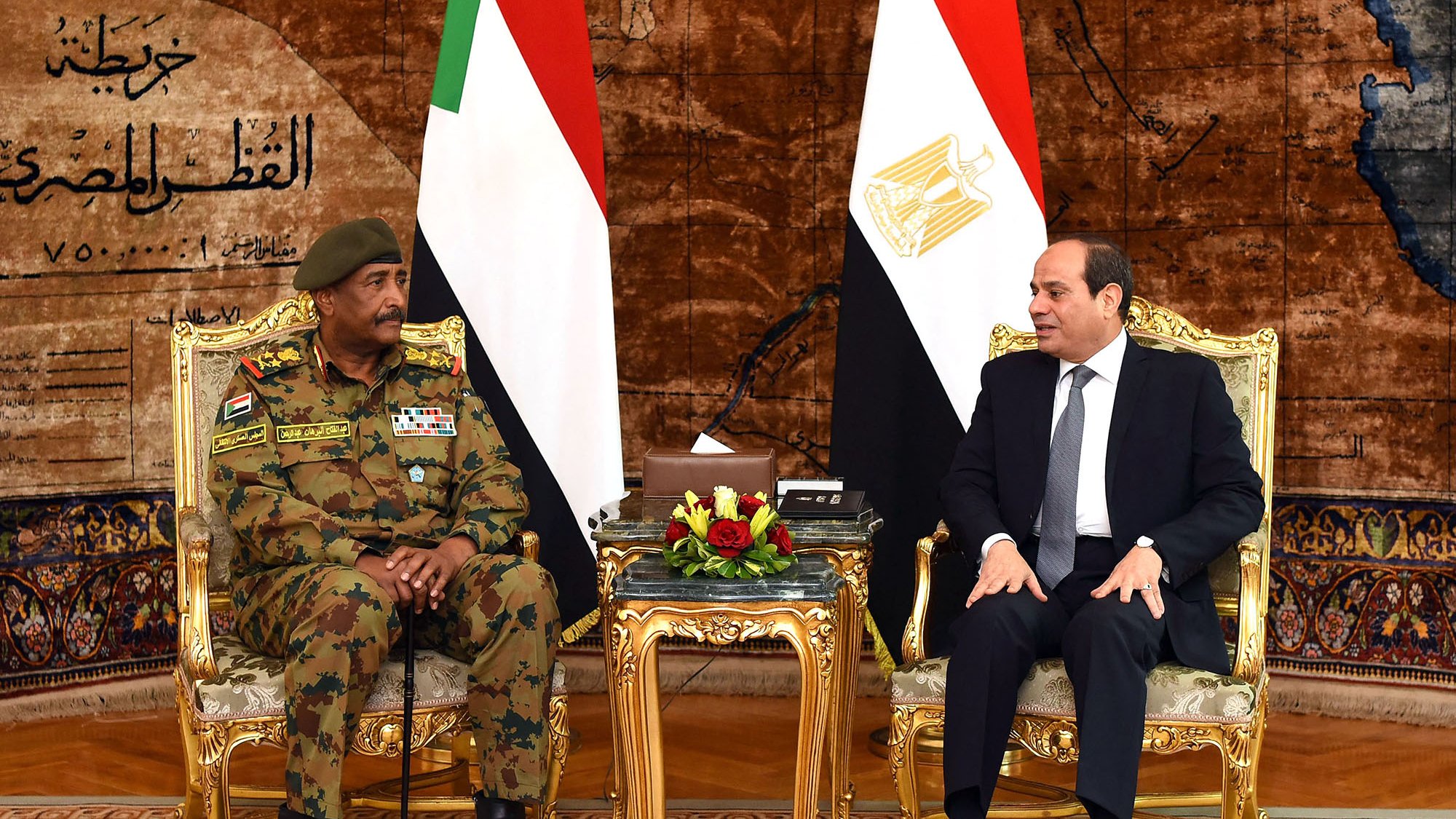 تظهر الصورة لقاءً رسميًا بين شخصين يجلسان على كراسي فاخرة أمام علمي السودان ومصر. خلفهم يوجد ستار مزخرف يضيف لمسة فخمة للمكان. الشخص على اليسار يرتدي زيًا عسكريًا ويبدو أنه مسؤول عسكري، بينما الشخص على اليمين يرتدي بدلة رسمية. يعبر كلا الشخصين عن تواصل دبلوماسي بين البلدين.