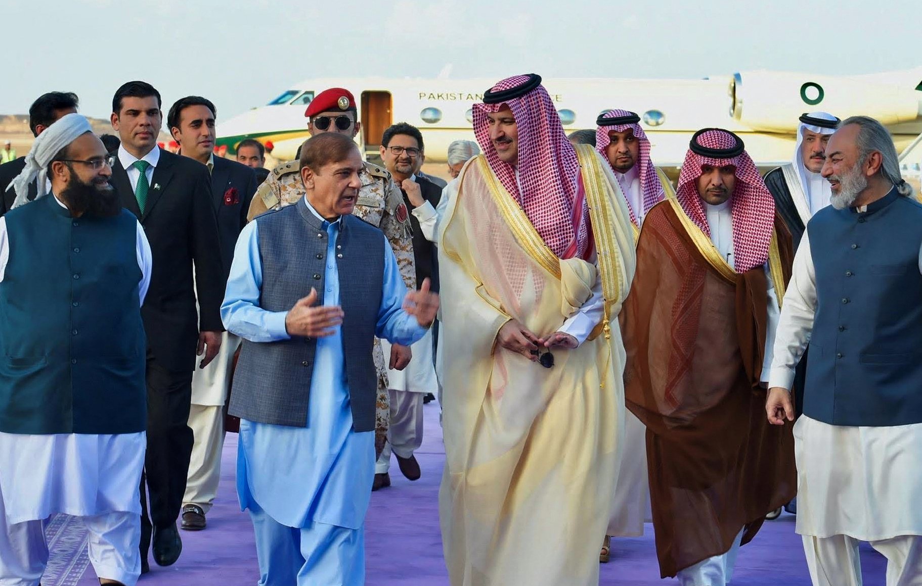 The image depicts a group of men walking together, likely during a formal event or diplomatic visit. They are dressed in traditional attire, with some wearing long robes and others in suits. The setting appears to be an airport or other official venue, indicated by the presence of a private jet in the background. The men seem engaged in conversation, and the atmosphere suggests camaraderie or collaboration. The purple carpet hints at a ceremonial welcome.