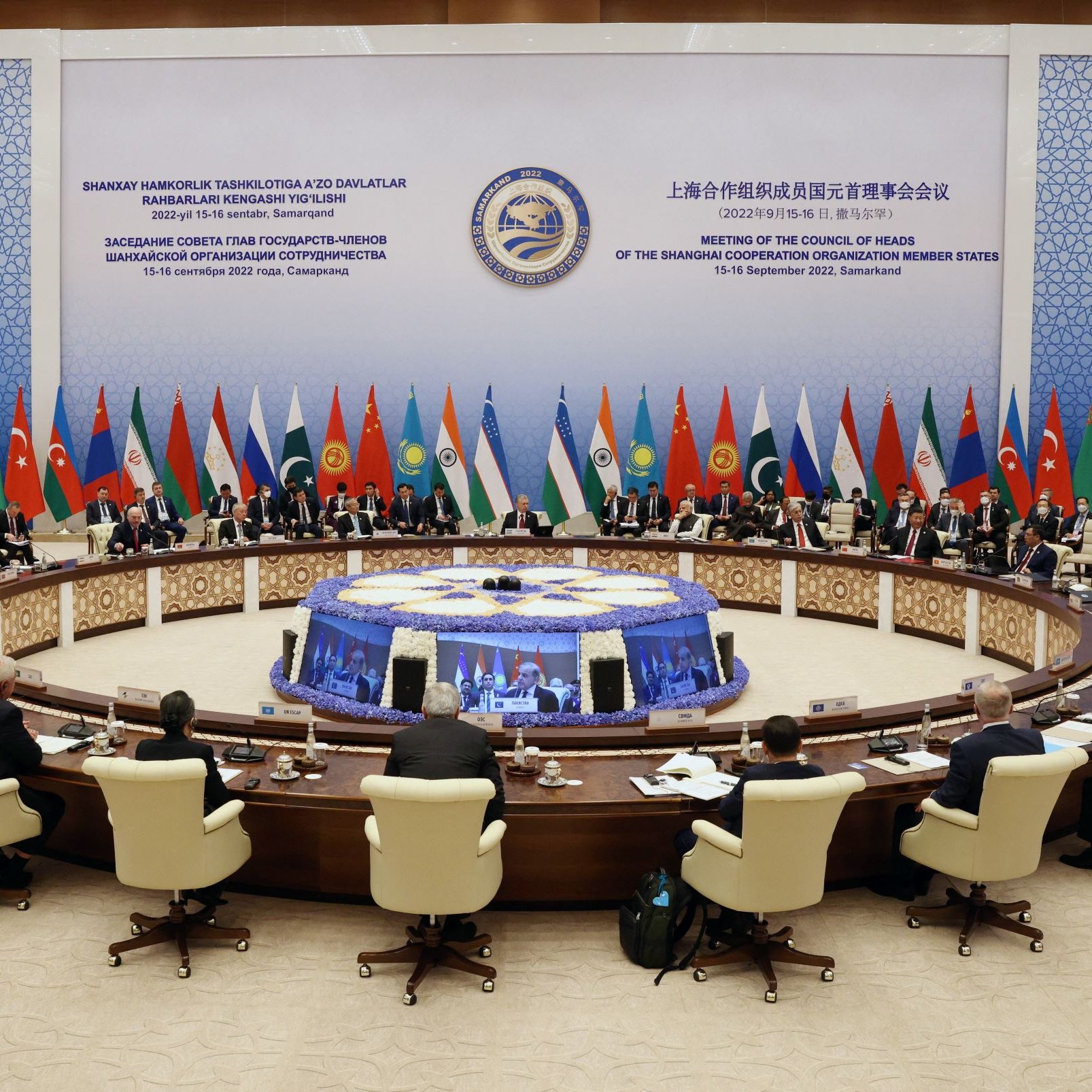 L'image montre une grande salle de conférence où se déroule une réunion. Des représentants de différents pays sont assis autour d'une table ovale, qui est ornée d'une décoration centrale. Les drapeaux de plusieurs nations sont visibles en arrière-plan, indiquant la diversité des participants. Les individus semblent engagés dans des discussions, et l'atmosphère est formelle. La pièce est bien éclairée et décorée de motifs qui évoquent un style moderne.
