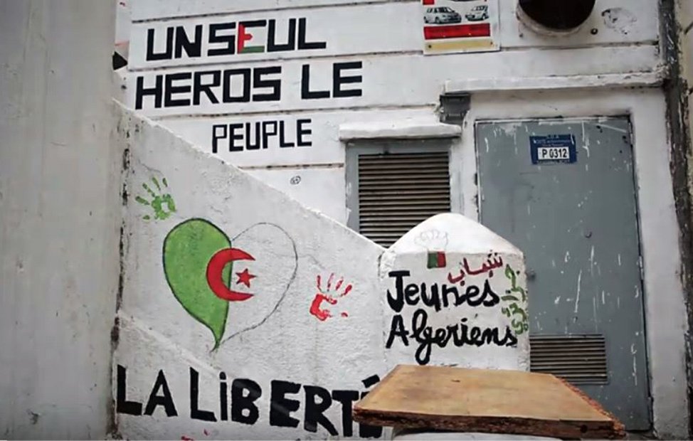 L'image montre un mur peint avec plusieurs graffitis. On y trouve des mots et des phrases exprimant des messages politiques et sociaux. Un dessin représentant un cœur dans les couleurs du drapeau algérien (rouge et vert) est visible. Il y a aussi une inscription en arabe, ainsi que des mots tels que "Jeunies Algériens" et "LA LIBERTÉ". Le fond suggère un lieu qui pourrait être un espace public, témoignant d'une expression artistique et militante.