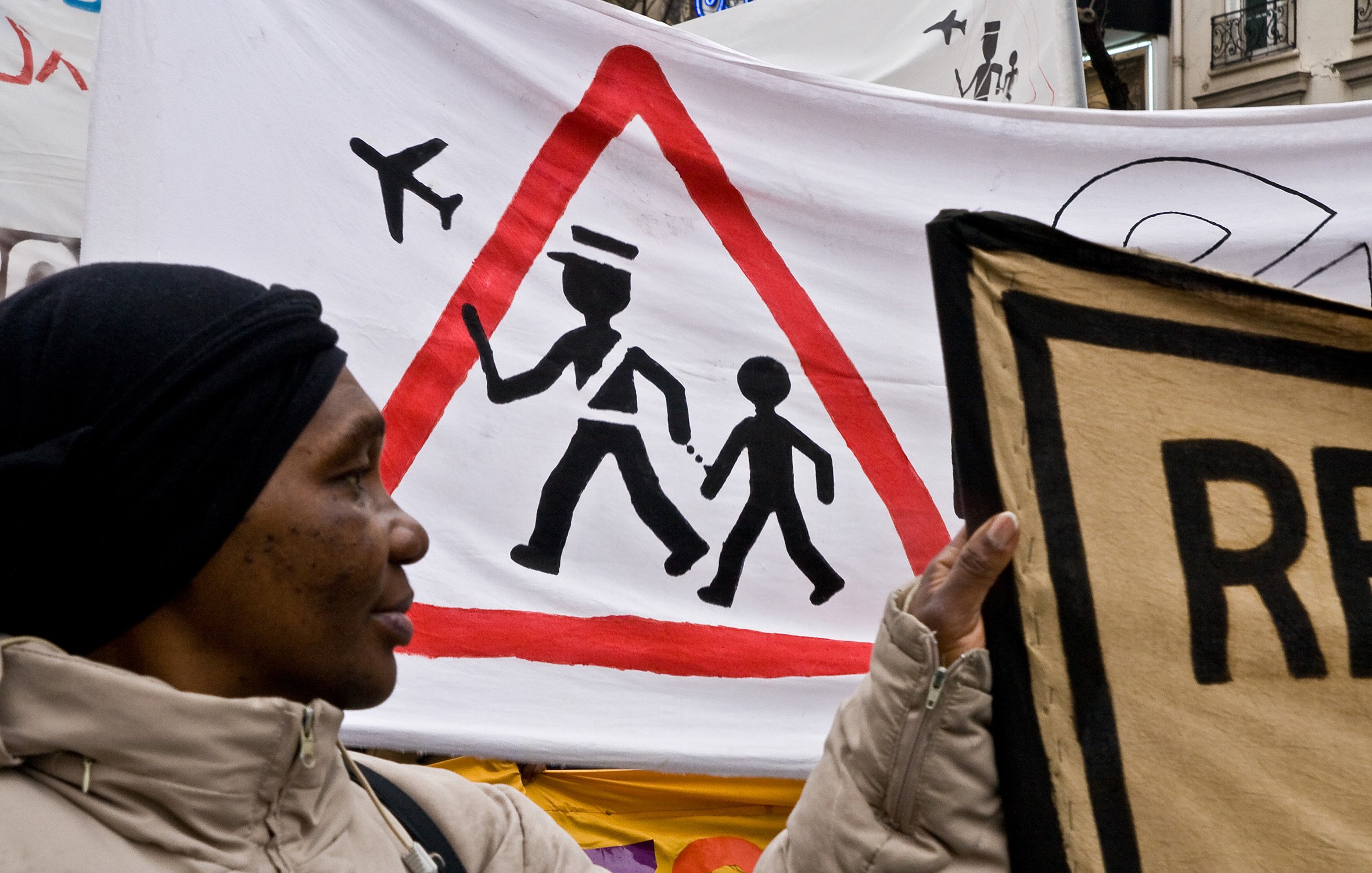 L'image montre une manifestation avec une femme tenant une pancarte. En arrière-plan, il y a une grande bannière avec un symbole d'avertissement en forme de triangle, représentant un adulte marchant avec un enfant. Ce symbole est accompagné d'illustrations d'avions en haut, suggérant une thématique liée au transport ou à la sécurité. La femme porte un manteau clair et un foulard noir, et son expression pourrait refléter l'engagement dans la cause qu'elle soutient.