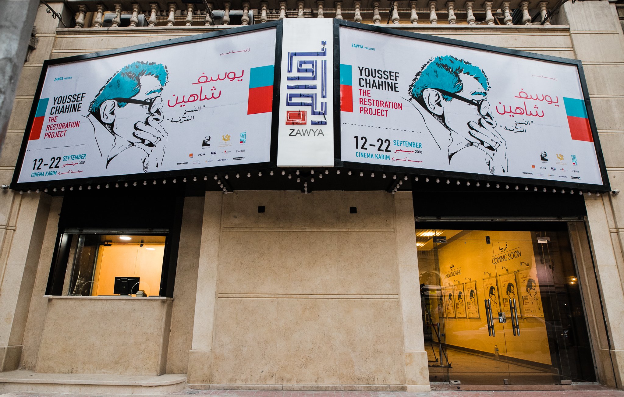 L'immagine mostra un edificio con una facciata decorata da grandi manifesti. I manifesti annunciano un progetto artistico intitolato "The Restoration Project" di Youssef Chahine, con date di evento indicate dal 12 al 22 settembre. Si può notare un design grafico accattivante, con illustrazioni e testo in arabo e in inglese. La zona sembra essere un luogo di cultura o un centro espositivo, con elementi architettonici tipici di un contesto urbano.