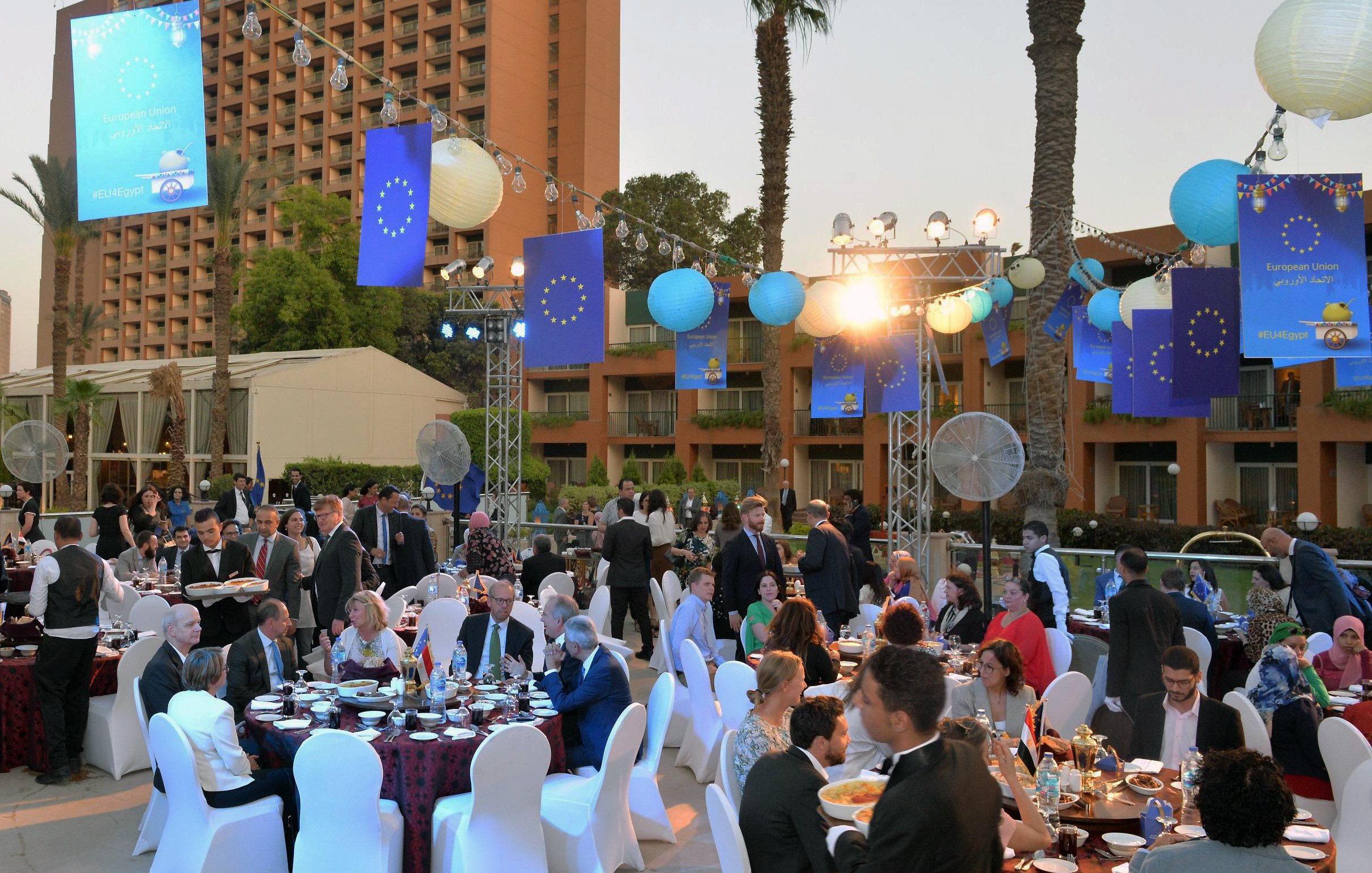 الصورة تظهر احتفالًا أو مناسبة اجتماعية في الهواء الطلق. هناك طاولات مزينة بأقمشة ملونة، وحولها مجموعة من الناس يرتدون ملابس أنيقة. في الخلفية، يبرز مبنى مرتفع، ويحيط بالمنطقة أشجار النخيل. الأضواء الداخلية والخارجية تعزز الأجواء الاحتفالية، وتوجد زينة على شكل كرات ملونة، بالإضافة إلى لافتات تحمل شعار الاتحاد الأوروبي. الأجواء تبدو مفعمة بالحياة والهدوء في الوقت نفسه.