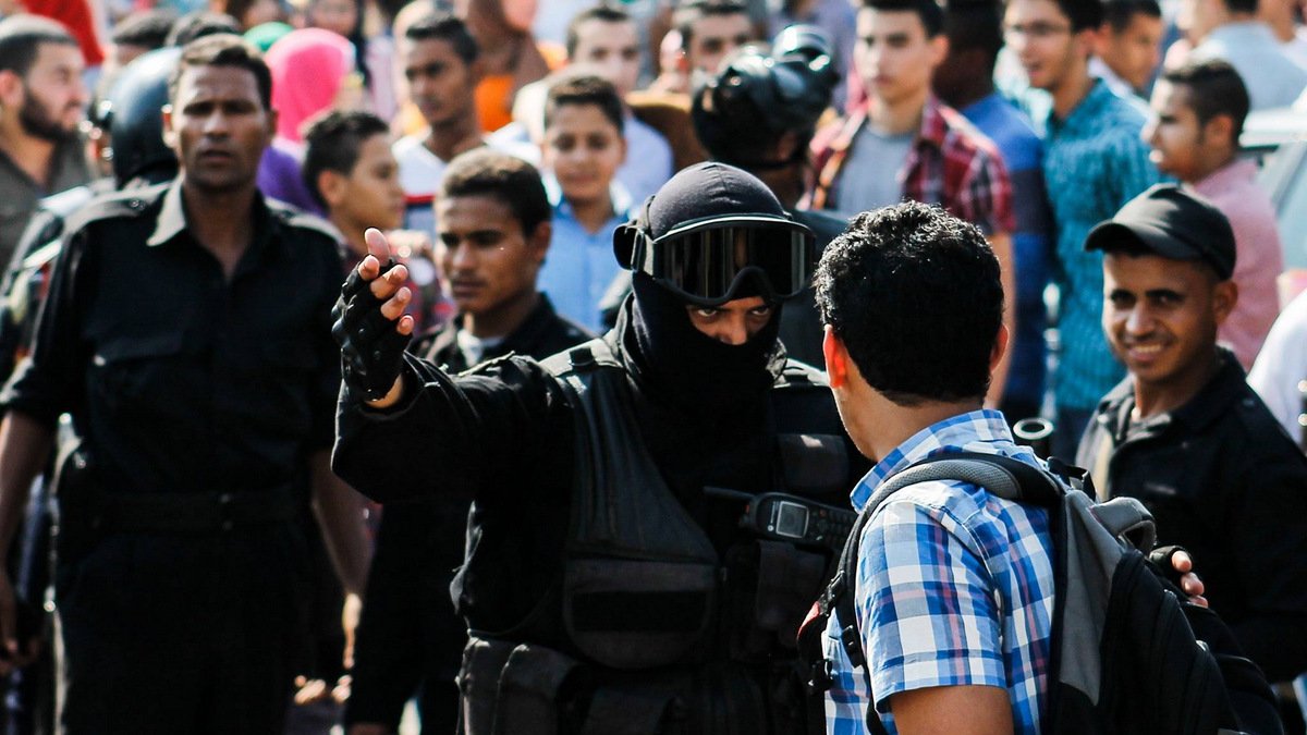 L'image montre une scène de confrontation dans un contexte urbain. À l'avant, un homme vêtu d'un uniforme noir, portant un casque et une visière, semble interagir avec un autre homme qui porte une chemise à carreaux. Ce dernier semble questionné ou arrêté, tandis qu'un groupe de personnes les entoure, observant la situation. On peut percevoir une atmosphère tendue, avec des expressions variées sur les visages des spectateurs. La scène suggère un moment de tension ou de dispute, probablement lié à une manifestation ou à une intervention policière.