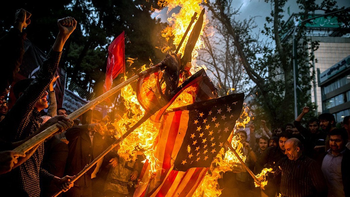 این تصویر نشان‌دهنده یک تجمع اعتراضی است که در آن گروهی از افراد در حال سوزاندن پرچم ایالات متحده هستند. در پس‌زمینه، دیده می‌شود که برخی از افراد دست‌ها را بالا برده و به نشانه اعتراض فریاد می‌زنند. دود و آتش در حال برخاستن از پرچم است و فضا به شدت احساسی و پر از شور و هیجان به نظر می‌رسد. درختان و ساختمان‌هایی نیز در پس‌زمینه وجود دارند که نشان‌دهنده مکان شهری این رویداد است.