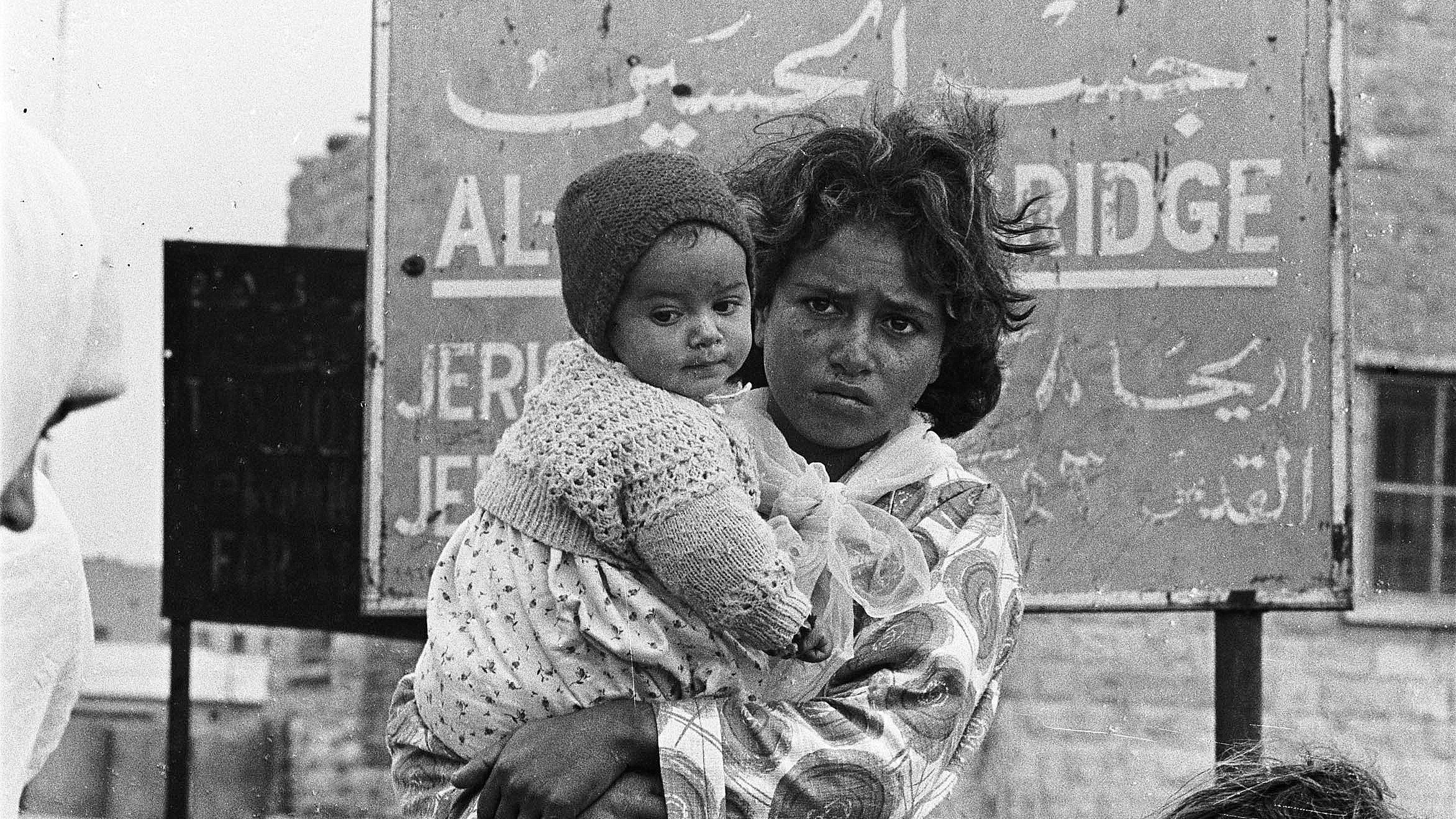 الصورة تُظهر فتاة صغيرة تحمل طفلة في حضنها. الفتاة تبدو متعبة ومرتبكة، تعبير وجهها يوحي بحالة من القلق أو الحزن. الطفلة ترتدي قبعة صوفية ولديها عيون واسعة. خلفهما يوجد لافتة مكتوبة باللغة العربية، تشير إلى مكان معين. الألوان في الصورة تبدو أحادية، مما يعكس أجواء تاريخية أو مأساوية.