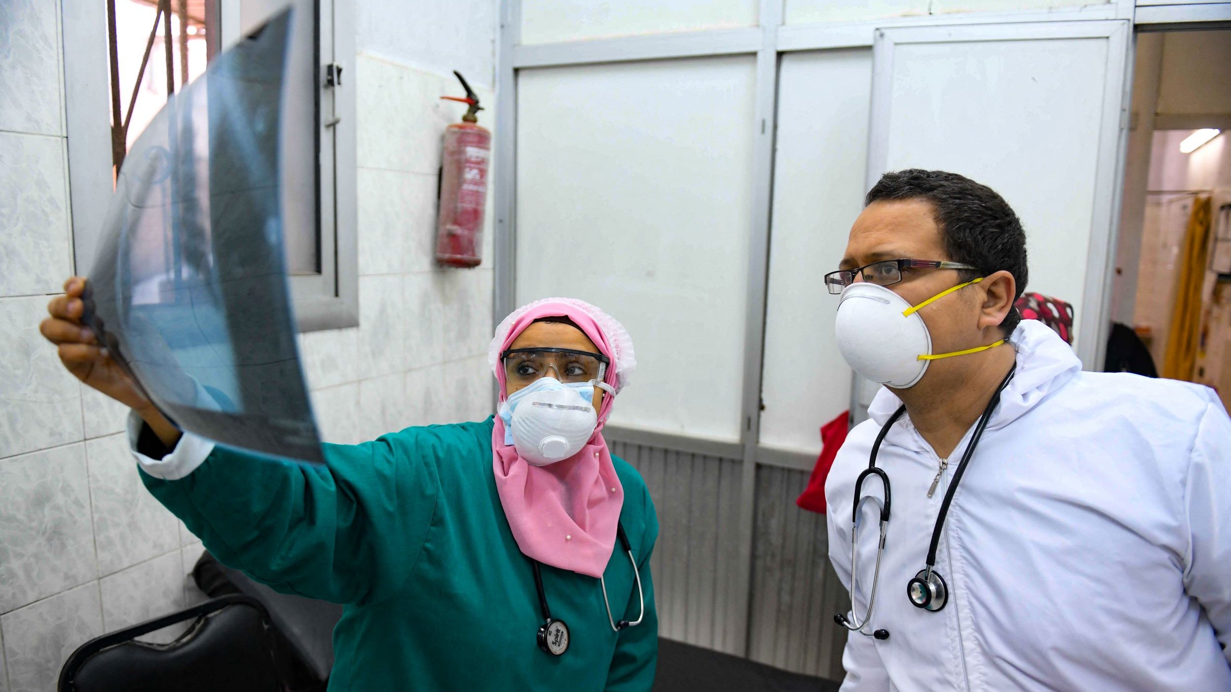 تظهر في الصورة مهنية طبية ترتدي زيًا طبيًا أخضر وغطاء رأس ورديًا، وتفحص صورة أشعة. إلى جانبها، يوجد طبيب يرتدي سترة بيضاء وقناع وجه وقناع تنفس. كلاهما يبدوان مهتمين بالمعلومات الموجودة في صورة الأشعة، ويظهر في الخلفية جدار مكتبي ومعدات طبية.