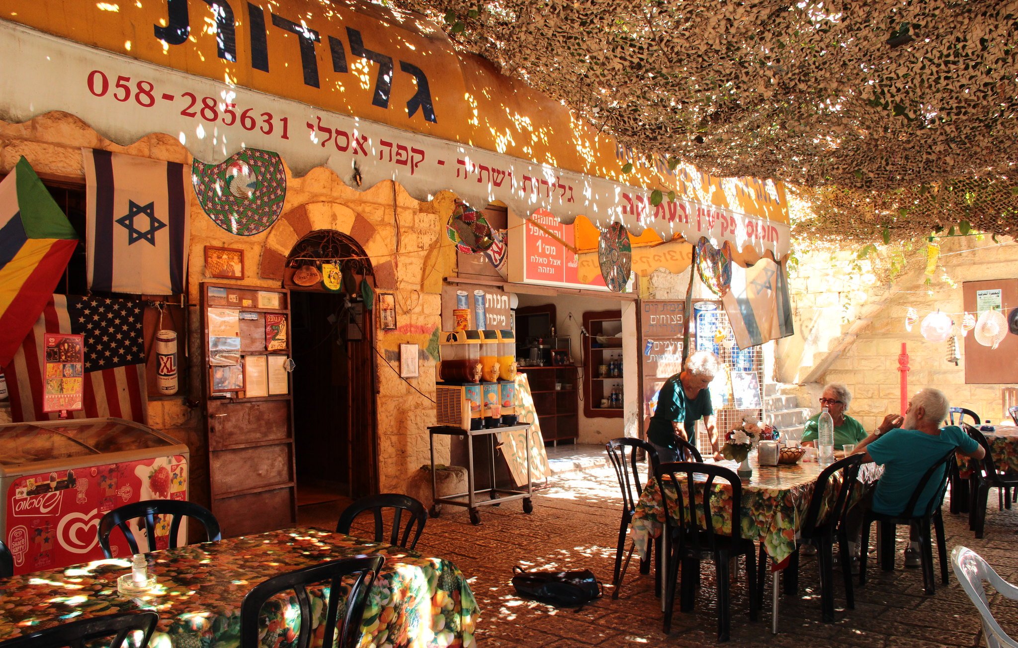 الصورة تظهر مقهى أو مطعمًا يحتوي على عدد من الطاولات والكراسي، ويبدو أن المكان مفتوح وتغطيه مظلة من الأوراق أو النباتات. الأجواء تبدو مريحة ومرحب بها، حيث يجلس عدد من الزبائن في أماكنهم. هناك لافتات باللغتين العبرية والإنجليزية، كما تظهر أعلام متنوعة، مما يدل على تنوع الزوار. يمتد المكان في منطقة جمجومية، مع ضوء الشمس يتسلل من خلال الأوراق.