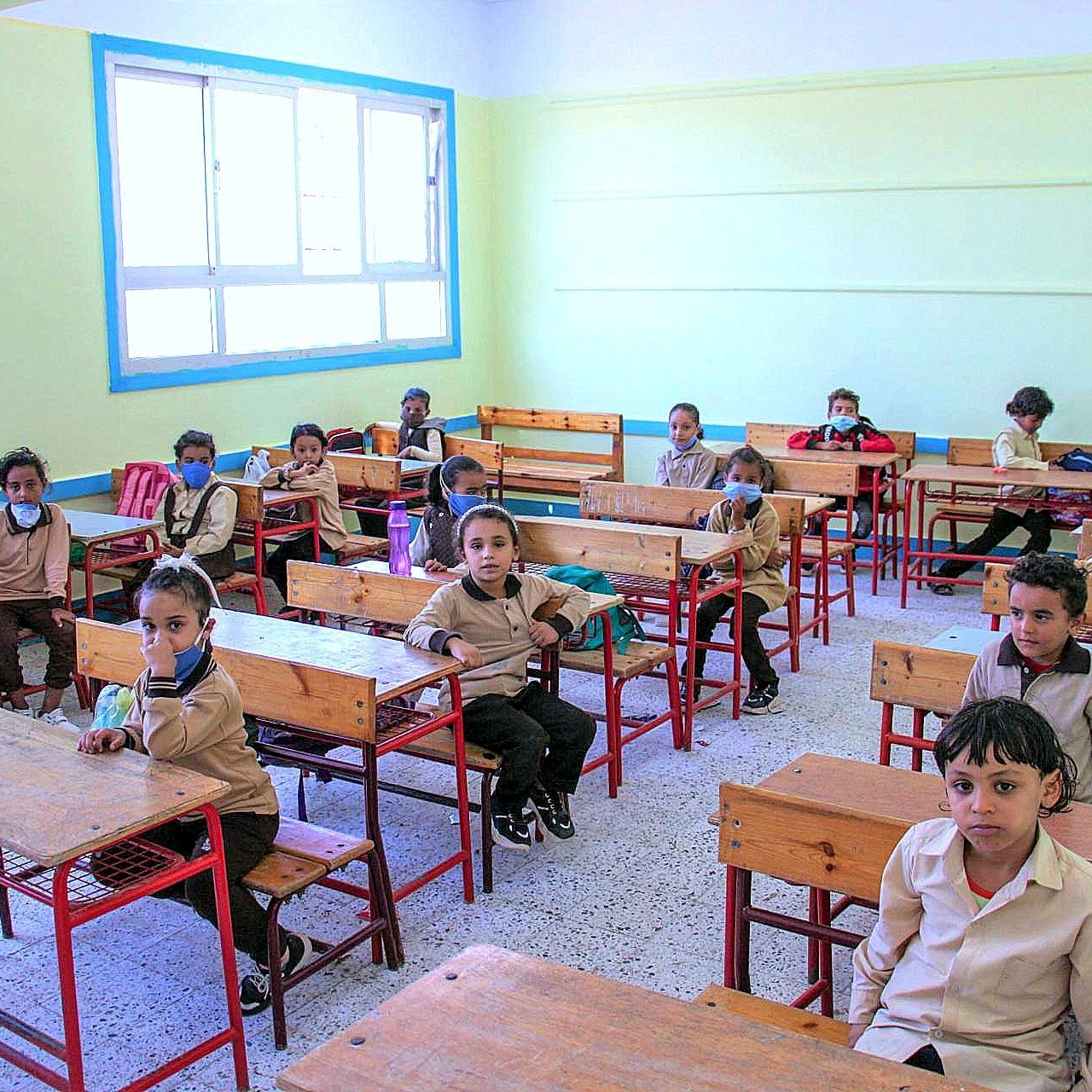 الصورة تُظهر فصلاً دراسياً يحتوي على عدد من الأطفال في مقاعدهم. الغرفة مضاءة جيداً بألوان فاتحة، الجدران مطلية بلون أخضر فاتح. الأطفال يرتدون زي المدرسة ويبدو عليهم الانتباه. بعضهم يرتدي كمامات. الطاولات والكراسي مصنوعة من الخشب. الغرفة منظمة ومرتبة وتحتوي على نوافذ كبيرة تسمح بدخول الضوء الطبيعي.