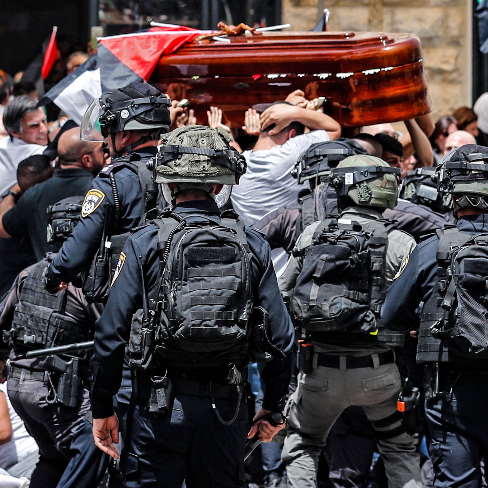 The image depicts a scene of tension and conflict. In the foreground, a group of police officers in tactical gear is facing a larger crowd of people who are mournfully carrying a wooden coffin. The crowd appears to be protesting or commemorating a significant event, with flags visible, including Palestinian flags. The atmosphere is charged, suggesting a clash between the police and the public. Emotions are high as both sides engage in this moment.