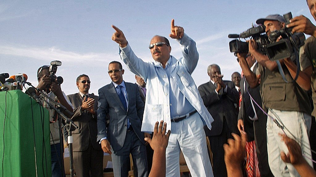 L'image montre un homme debout sur une scène, habillé en costume clair. Il semble être en train de faire un discours et de pointer vers le public. Autour de lui, plusieurs personnes l'écoutent attentivement, dont certaines sont en train de prendre des photos ou de filmer avec des caméras. Il y a une ambiance d'événement public ou politique, avec un discours énergique et engageant.