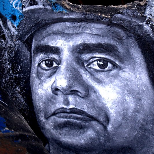 تظهر الصورة رسما جداريًا لوجه رجل، حيث يتميز التعبير بالعمق والتفاصيل الدقيقة. يُظهر الرسم عيونًا تعكس مشاعر قوية، والخطوط تتباين بين الظل والنور، مما يعطي إحساسًا بالحياة والواقعية. الخلفية تحتوي على ألوان داكنة ومخططات فنية أخرى، مما يبرز التركيز على وجه الرجل ويعزز من جمال العمل الفني.