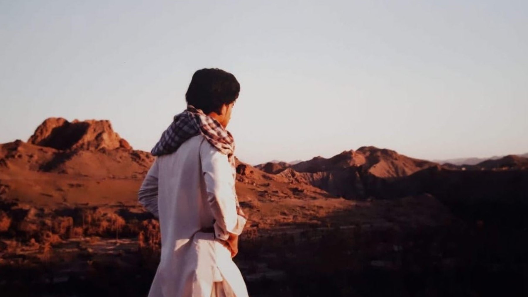L'image montre une personne debout sur une colline, regardant au loin vers un paysage montagneux. La lumière douce du coucher de soleil illumine les montagnes environnantes, créant une atmosphère sereine. La personne porte une tenue traditionnelle, avec un chèche sur la tête, qui évoque une culture spécifique. L'arrière-plan est composé de collines et de paysages arides, typiques de certaines régions désertiques.