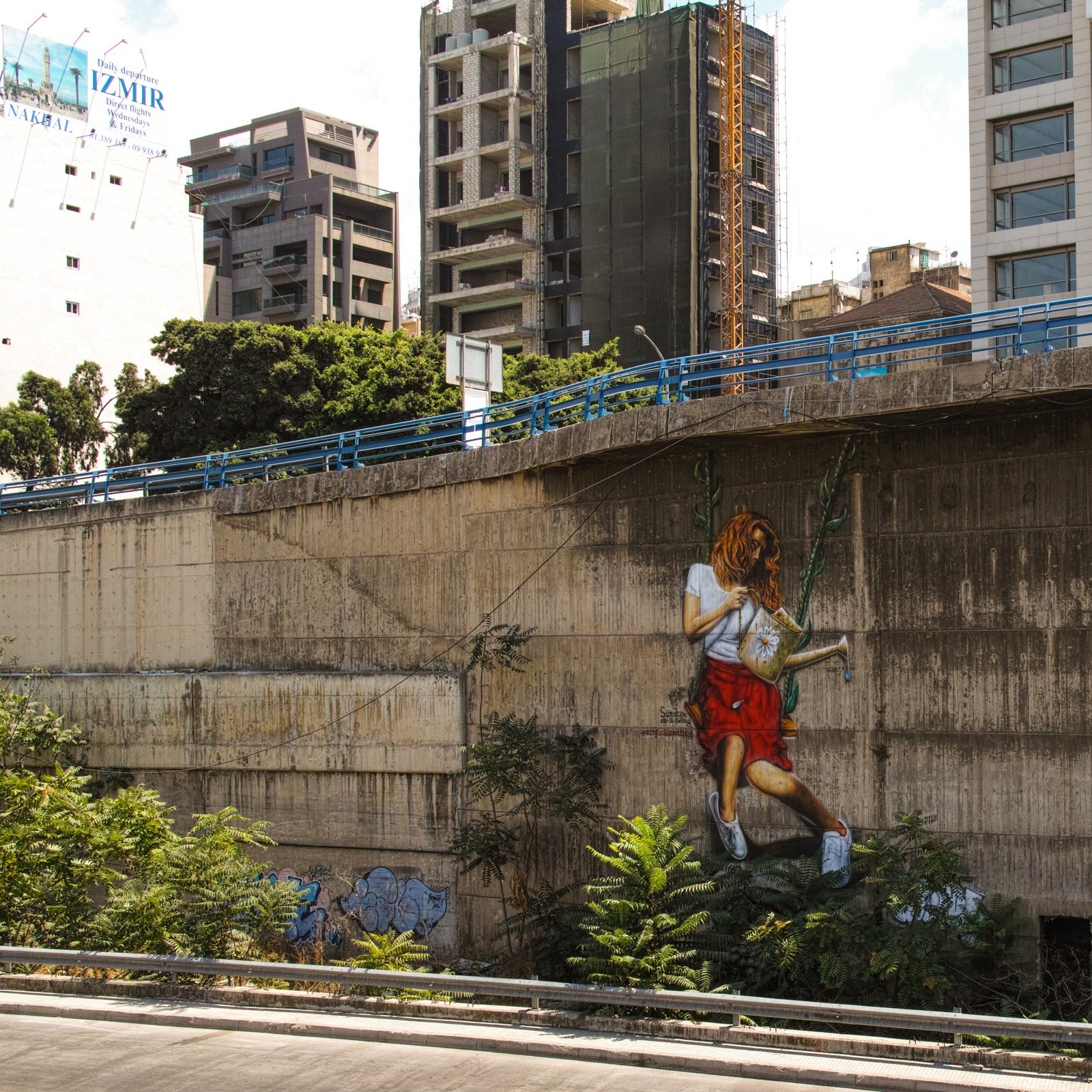 L'image montre un mur urbain en béton, partiellement recouvert de végétation. À droite, une grande peinture murale représente une personne en train de jouer à la balançoire, accrochée au mur. En arrière-plan, on aperçoit des bâtiments modernes, certains en construction, ce qui donne une ambiance de ville dynamique. La scène mélange l'art urbain avec des éléments naturels, créant un contraste intéressant entre le béton et la verdure.