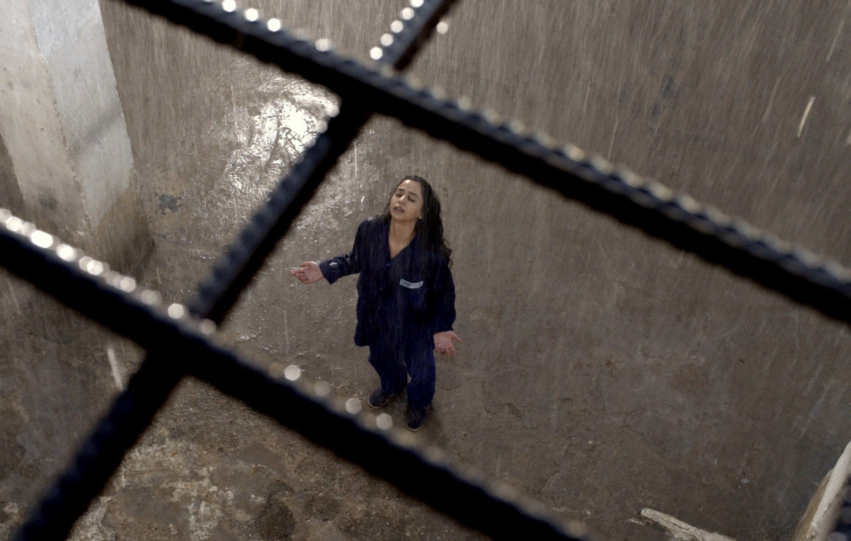 L'image montre une scène dramatique d'une femme debout dans un espace clos, probablement une cellule. Elle porte une tenue de prison et semble être sous une forte pluie qui tombe du plafond. La lumière est tamisée, créant une ambiance sombre et mélancolique. La femme, les bras ouverts et la tête inclinée vers le ciel, exprime une pose de désespoir ou de lâcher-prise, tandis que des barreaux en fer témoignent de son confinement. Le sol semble humide et dégradé, ce qui renforce l'atmosphère oppressante de l'environnement.