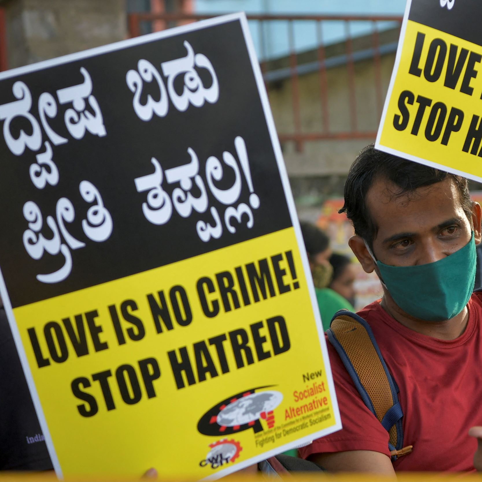 The image depicts a person holding two large signs that read "LOVE IS NO CRIME! STOP HATRED!" in both English and Kannada, a language spoken in India. The individual is wearing a face mask and appears to be participating in a demonstration or rally, advocating for love and against hate. The atmosphere suggests a sense of activism and a call for social change, with a crowd visible in the background. The signs are bright and eye-catching, emphasizing the message strongly.
