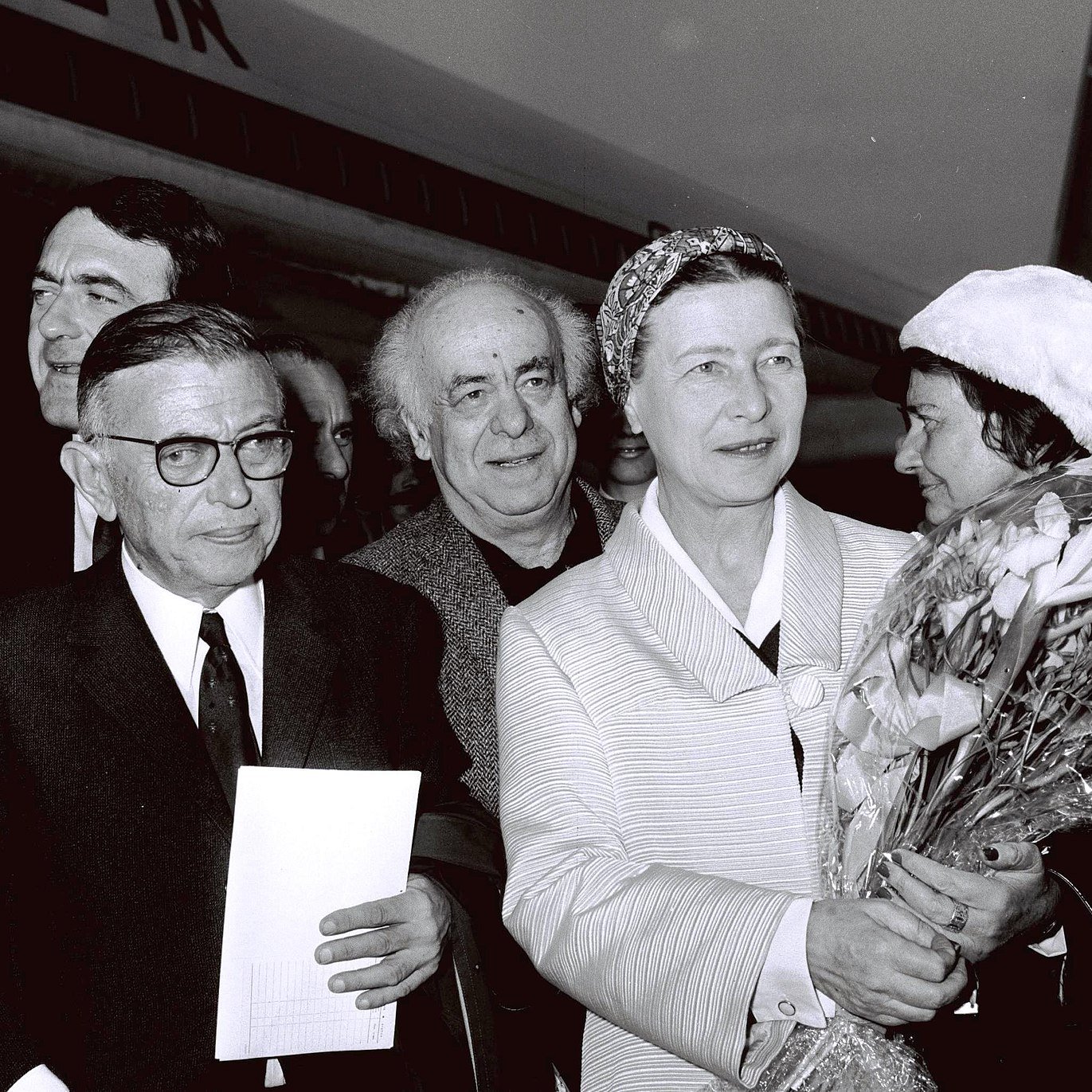 The image captures a group of individuals outside an airplane, likely at an airport. There are six people in the frame, with one woman prominently holding a bouquet of flowers. She appears to be the center of attention, dressed in a light-colored outfit and wearing a headscarf. The men around her are dressed in suits and vary in expressions. The background includes the tail of the aircraft, suggesting a public or ceremonial occasion. The image is in black and white, giving it a classic, historical feel.