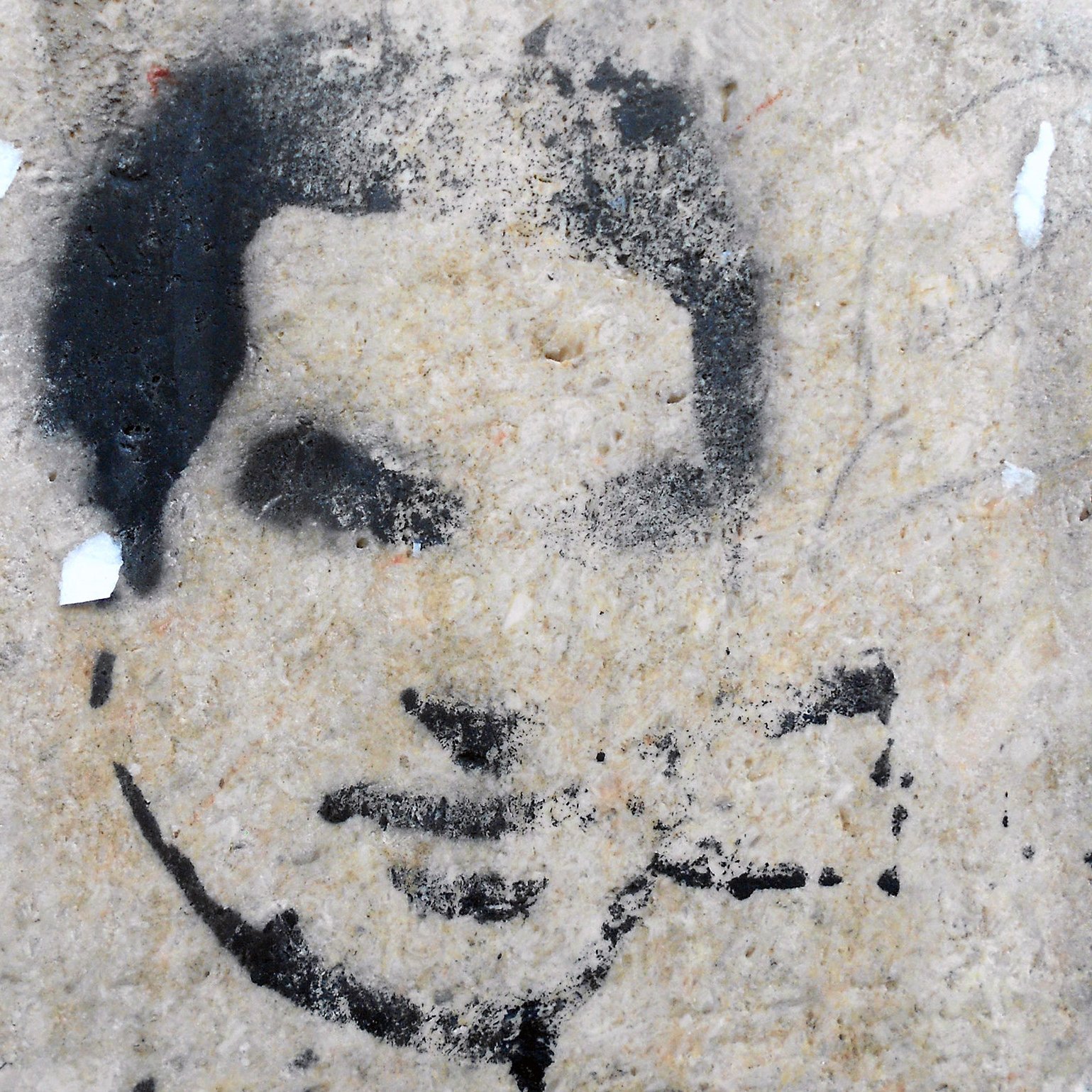 این تصویر شامل یک طرح یا گرافیتی روی دیوار است که چهره‌ای را به صورت سیاه و سفید نشان می‌دهد. جزئیات چهره به وضوح قابل تشخیص نیستند و بیشتر به صورت سایه‌ای به نظر می‌رسند. زمینه تصویر ممکن است از جنس بتن یا دیوار سنگی باشد که به مرور زمان دچار wear و تخریب شده است. 