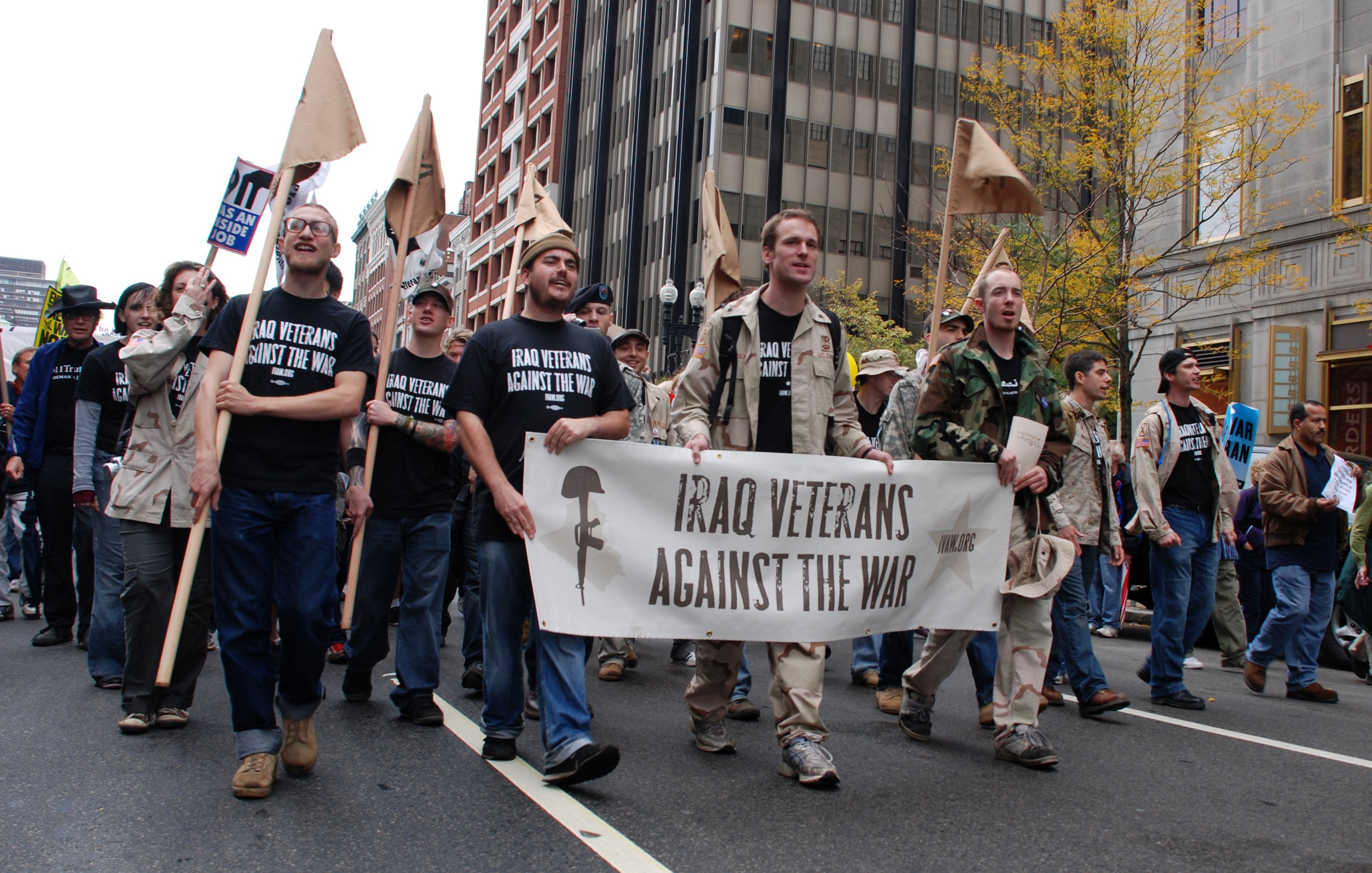 L'image montre un groupe de manifestants marchant dans une rue, tenant des pancartes. Au premier plan, plusieurs personnes portent des t-shirts noirs avec des slogans, et l'un d'eux tient une grande banderole qui dit "Iraq Veterans Against The War". Il semble s'agir d'une manifestation contre la guerre en Irak, avec des participants d'âges et d'apparences variés. L'ambiance semble sérieuse et engagée. Des bâtiments urbains et des arbres aux feuilles jaunes sont visibles en arrière-plan, suggérant que la scène se déroule en automne.