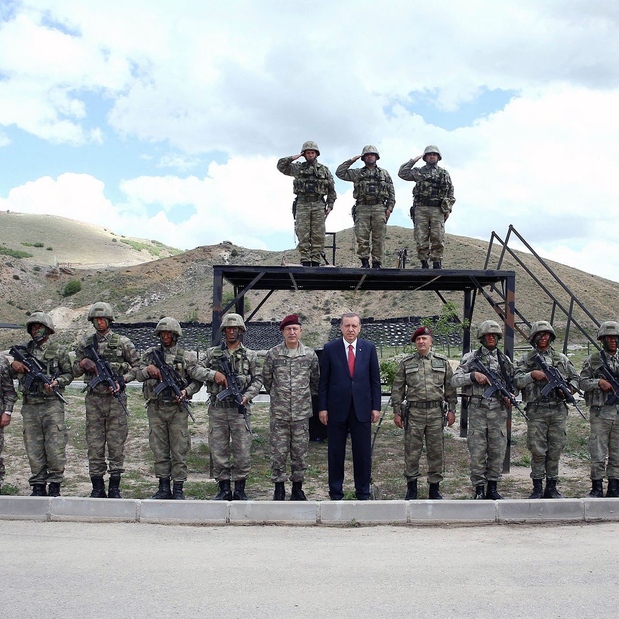 La imagen muestra a un grupo de soldados en un entorno militar. Hay un total de quince soldados alineados, vestindo uniformes de camuflaje y sosteniendo sus armas. En el centro de la imagen, se encuentra un hombre vestido con traje, aparentemente en una posición de liderazgo. Al fondo, se pueden ver dos soldados más en una plataforma elevada, saludando en posición de firme. El paisaje que rodea al grupo es montañoso y despejado, con nubes en el cielo.