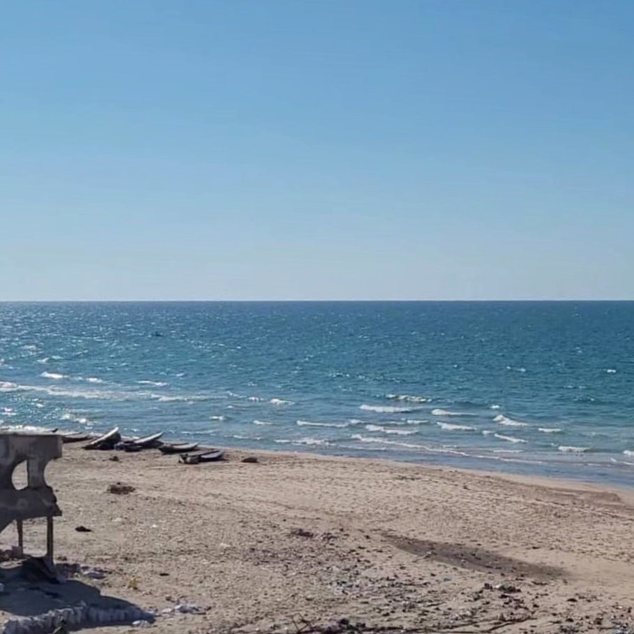 L'immagine mostra una spiaggia con sabbia chiara e un mare blu intenso sotto un cielo sereno e azzurro. In lontananza si possono vedere alcune imbarcazioni sulla superficie dell'acqua. Una struttura diroccata, grigia e in parte crollata, si trova sulla spiaggia, creando un contrasto con il paesaggio naturale circostante.
