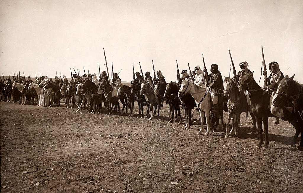 این تصویر نشان‌دهنده گروهی از سواران را در یک خط منظم است. آن‌ها بر روی اسب‌های خود نشسته‌اند و برخی از آن‌ها نیز نیزه یا سلاح‌هایی در دست دارند. لباس‌های آن‌ها سنتی به نظر می‌رسد و فضایی تاریخی را القا می‌کند. زمین زیر پای آن‌ها خشک و سنگی است و در پس‌زمینه، فضای باز و آسمان دیده می‌شود. این تصویر می‌تواند اشاره به یک دوره خاص از تاریخ یا مراسمی فرهنگی داشته باشد.